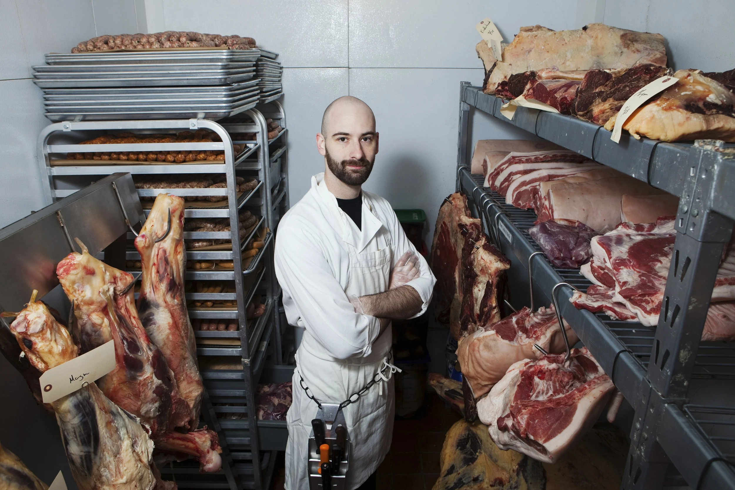 A butcher in a meat storage room with racks of various cuts of raw meat and sausage logs around him.