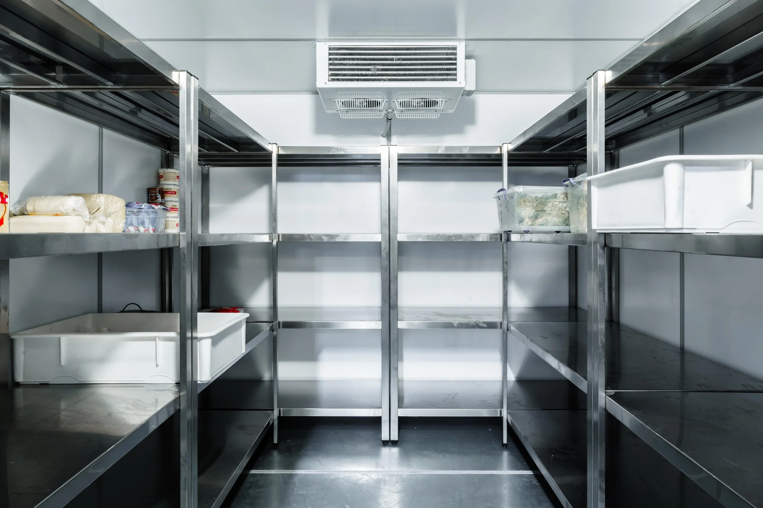 Empty stainless steel storage shelves in a cold storage room or walk-in freezer, with a mounted air conditioning unit on the ceiling.