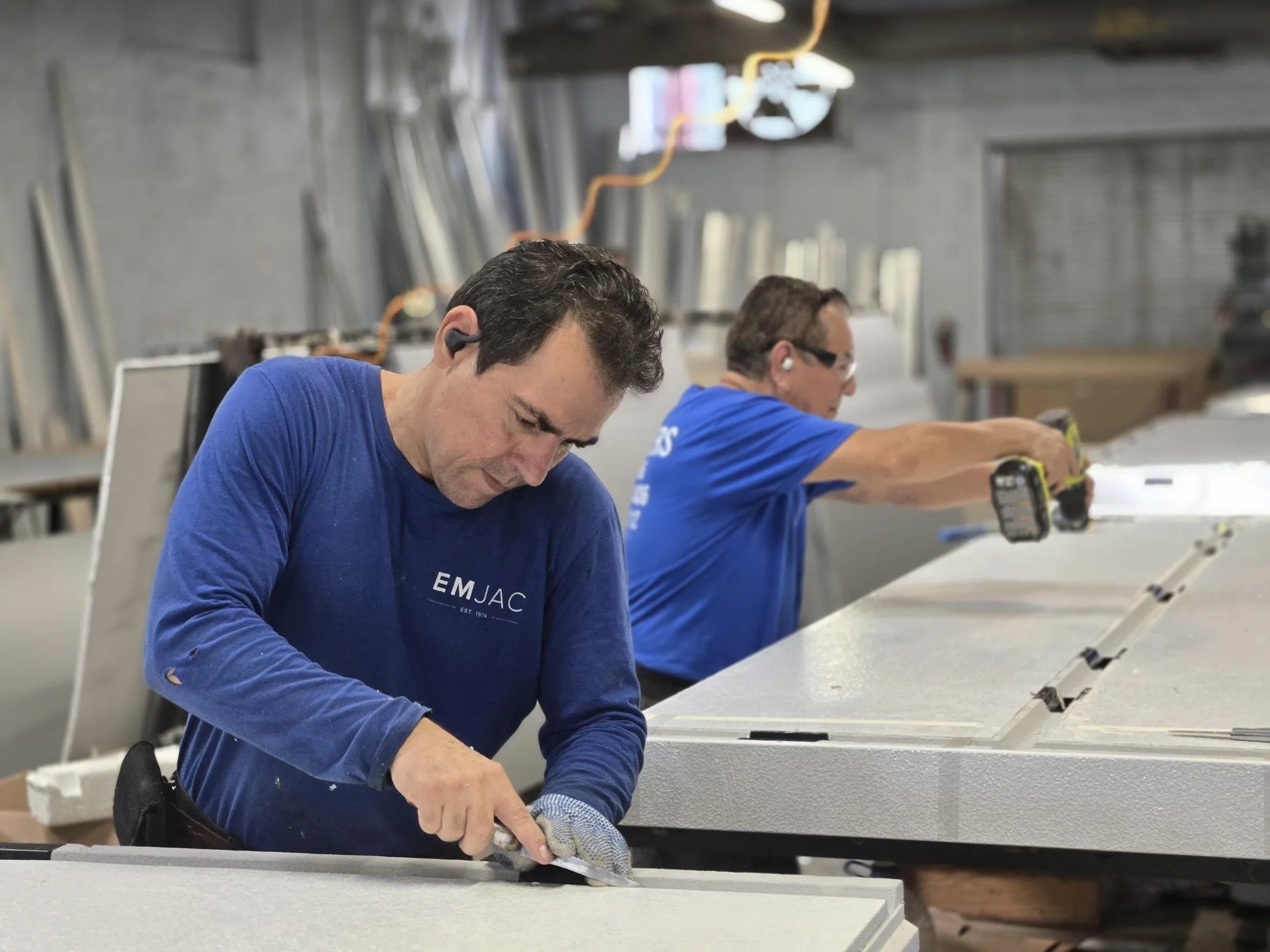 Two men working on a large piece of material in a workshop, using power tools, wearing safety glasses and at workbenches.