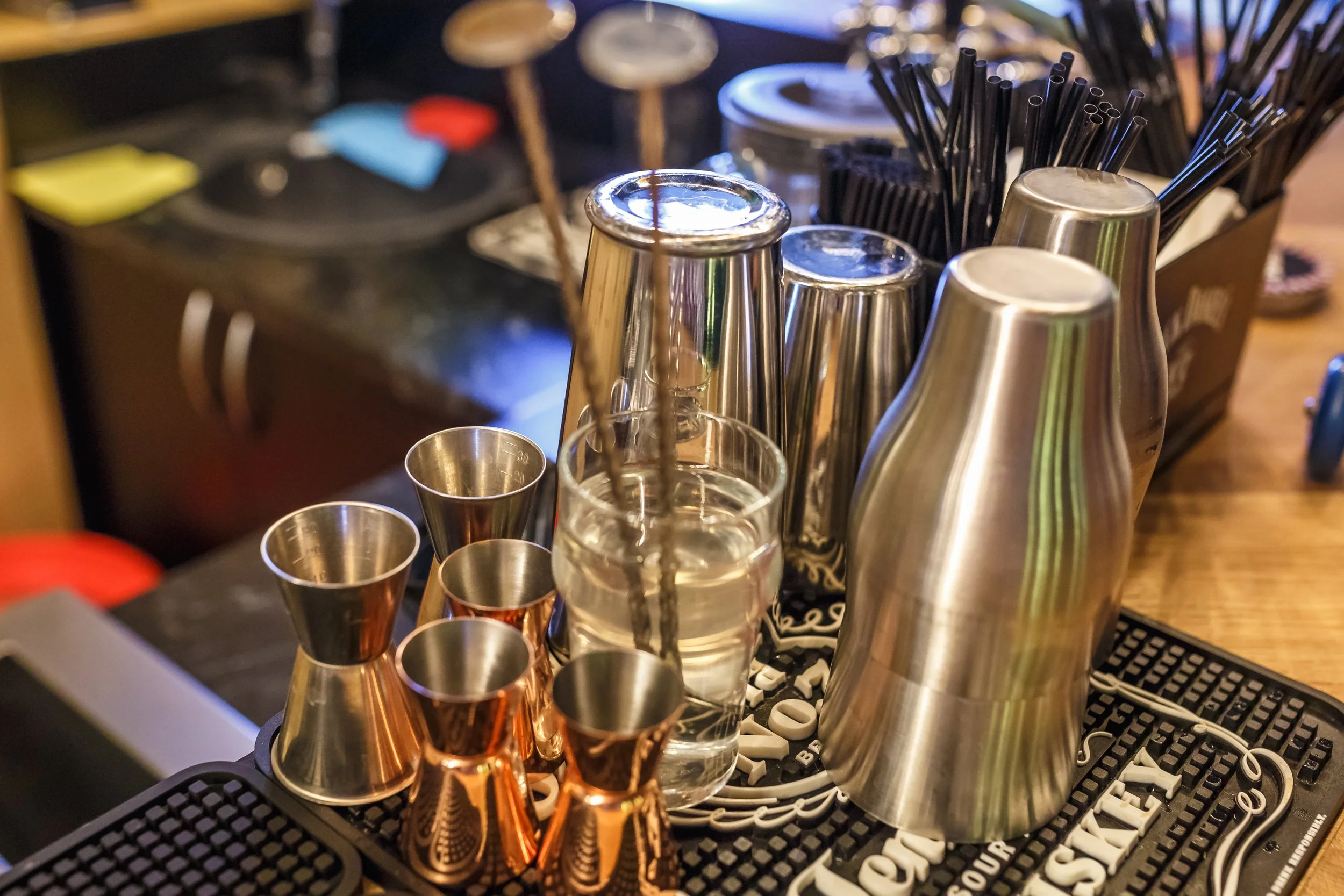 Bar countertop with metal cocktail shakers, shot glasses, a glass of water, black straws, and bar tools, with a cash register in the background.