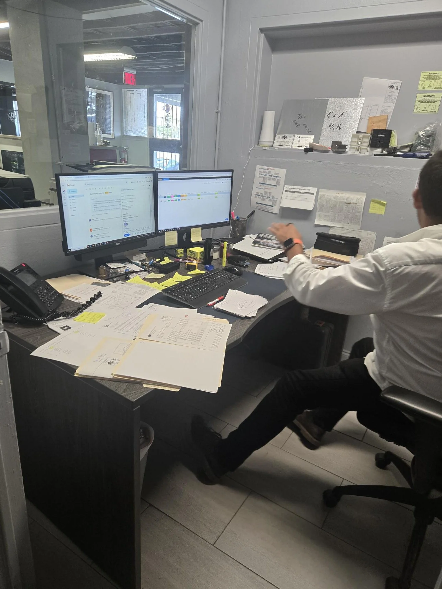 Office desk cluttered with papers, computer monitors, and office supplies, with a person sitting at the desk facing away.