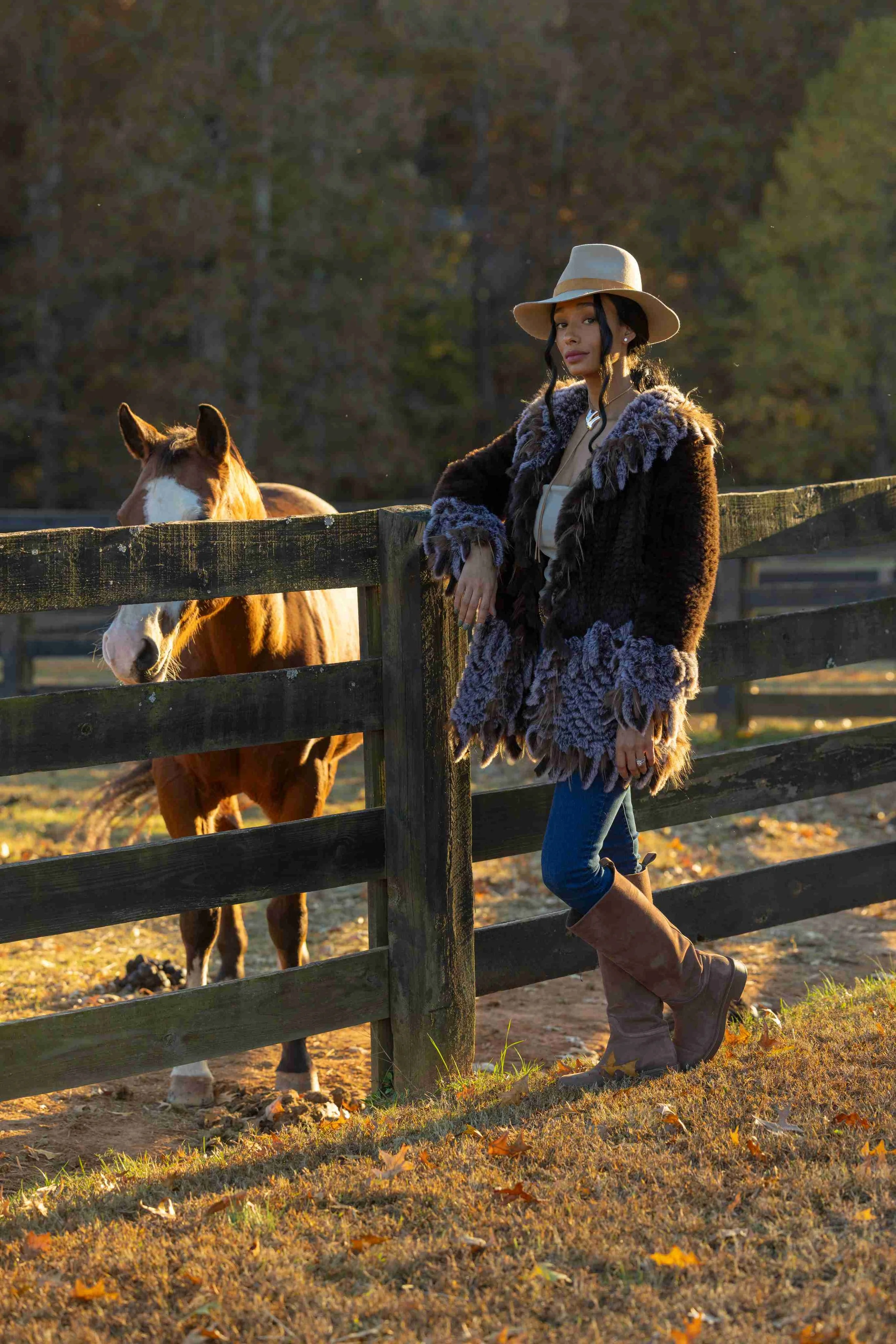 A woman leaning on a wooden fence with a horse beside her in an outdoor setting during sunset, with trees in the background and fallen autumn leaves on the ground.