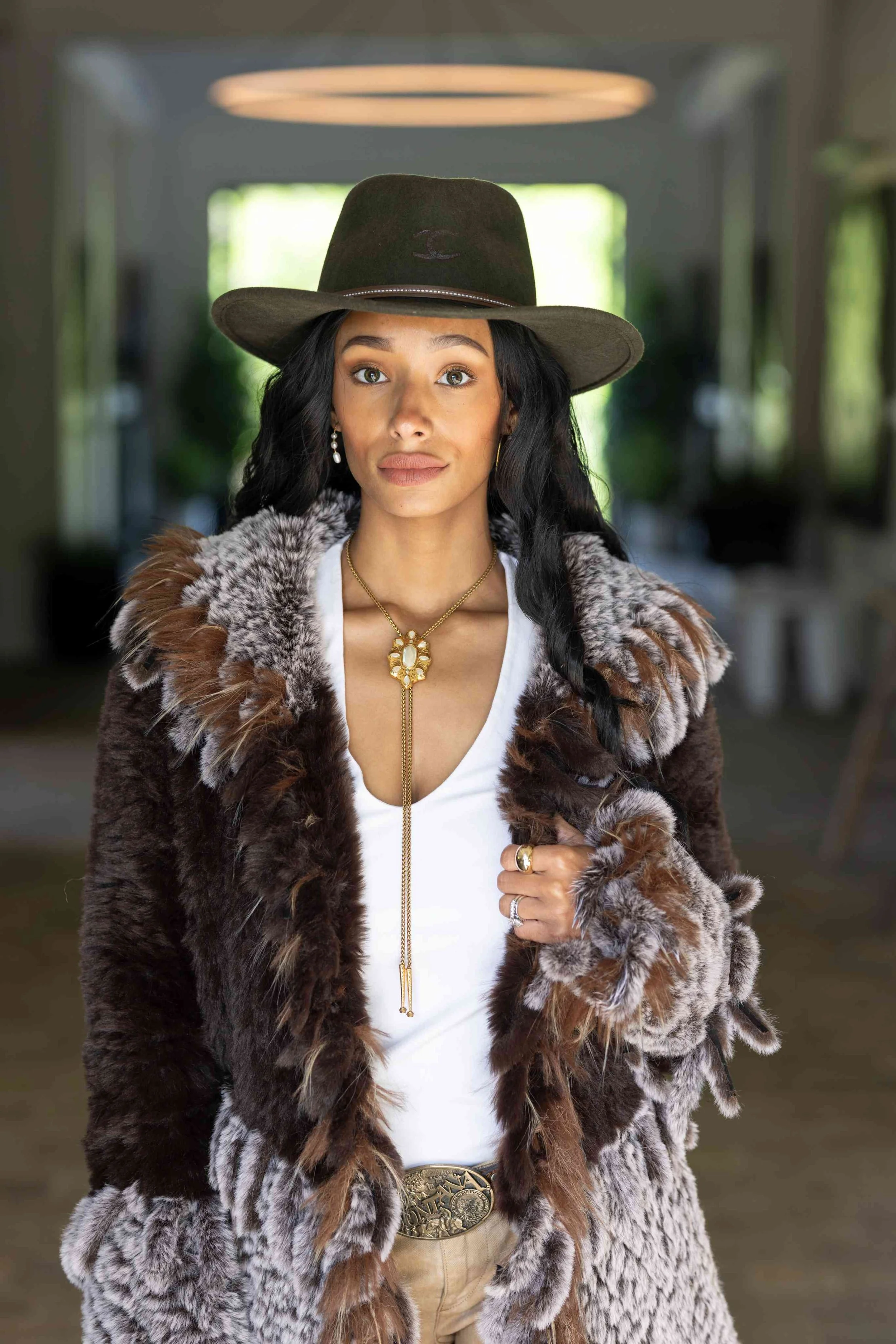 A woman with long black hair wearing a wide-brimmed hat, gold jewelry, a fur coat, and a white top, standing in an indoor setting with blurred background.