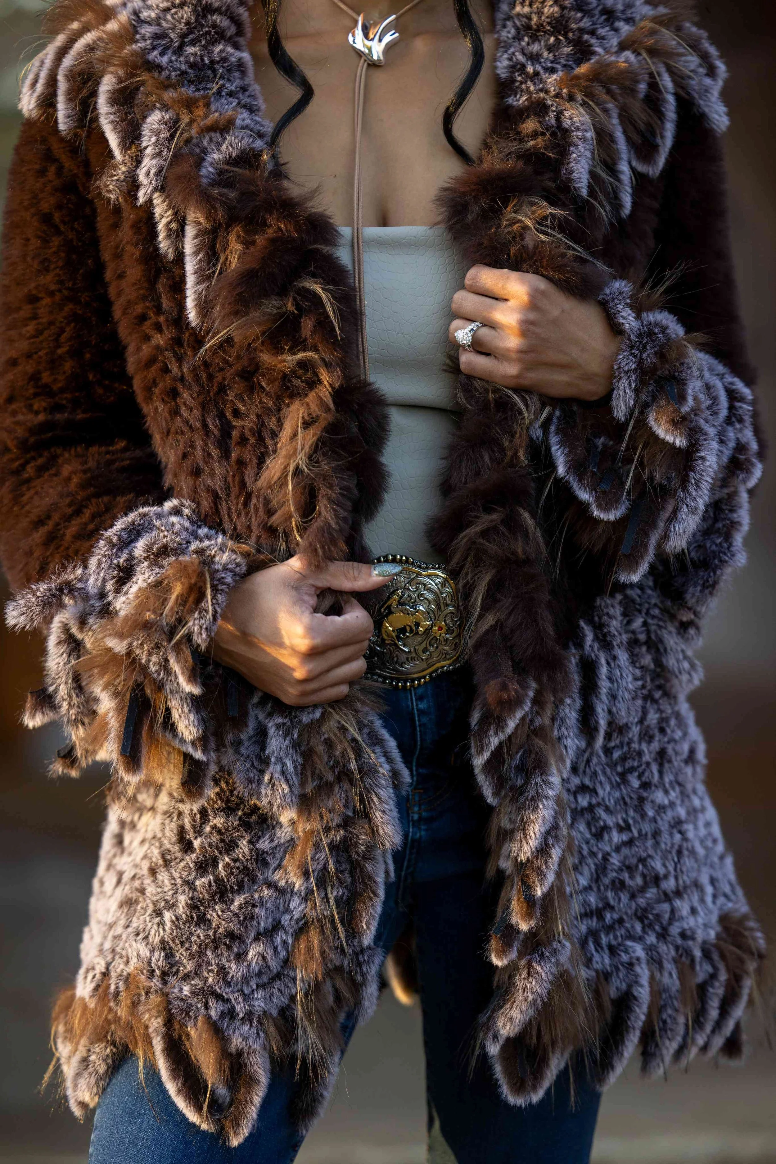 Person wearing a large, plush fur coat with brown, black, and gray tones, holding a belt with a decorative buckle, and wearing a ring with a large stone. The person's face is not visible.