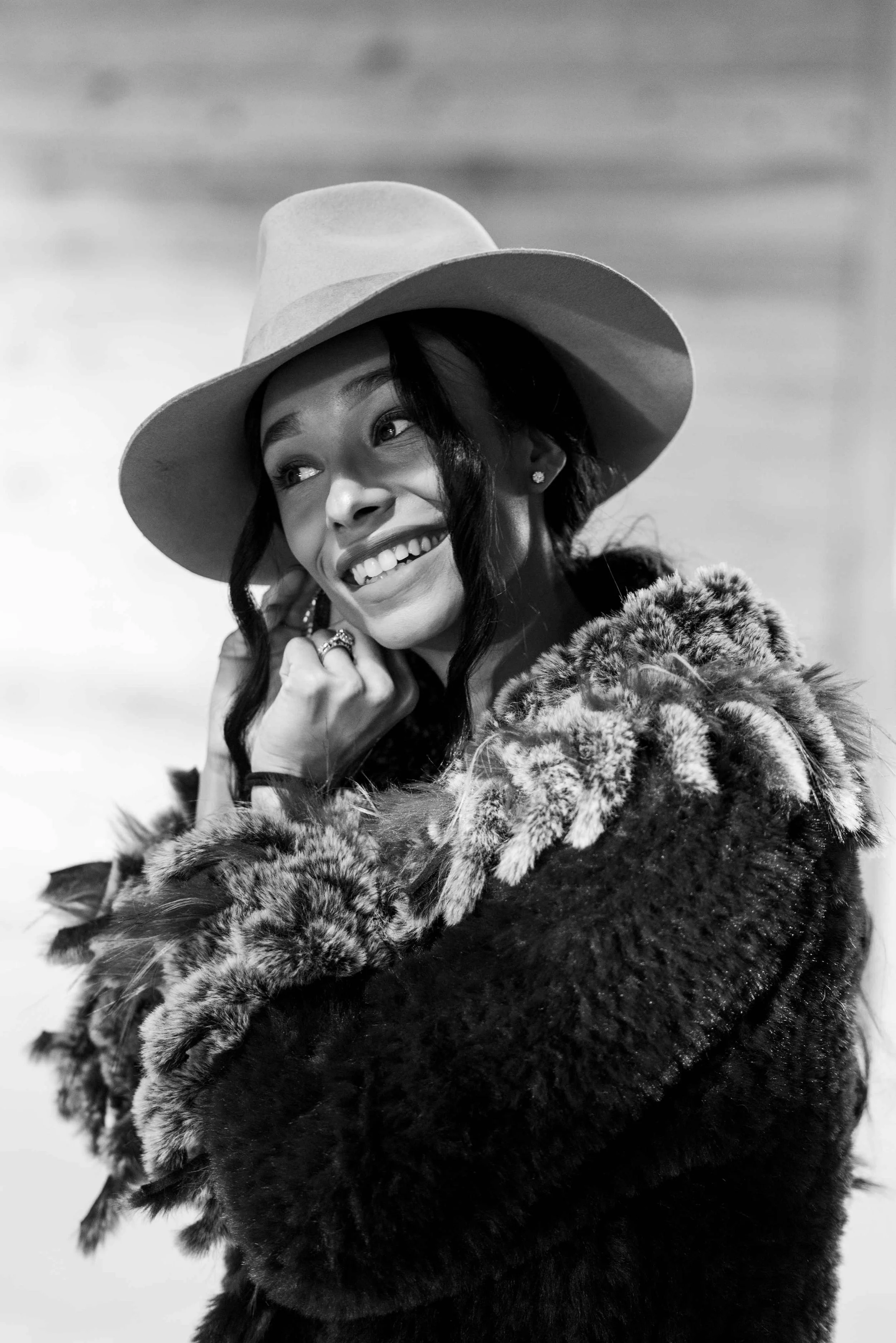 A smiling woman wearing a wide-brimmed hat, earrings, and a fur coat, with a cloudy sky background.