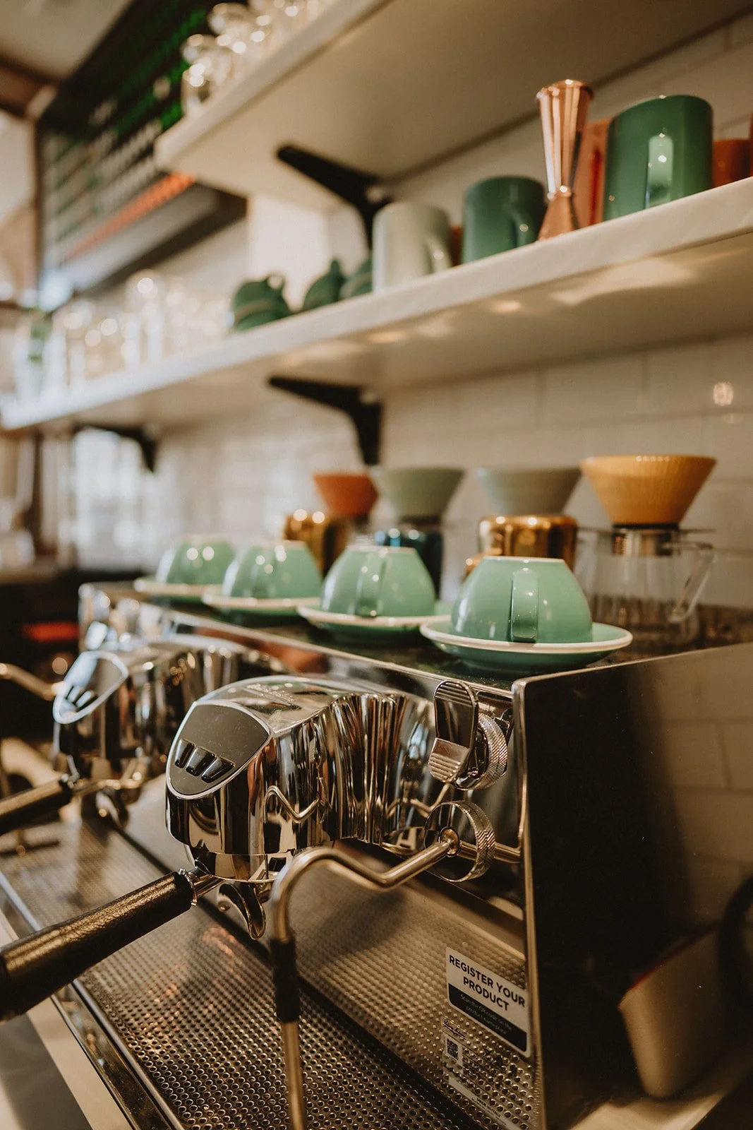 Close-up of a stainless steel espresso machine with green and brown cups, bowls, and a cold brew glass on shelves behind it in a coffee shop.