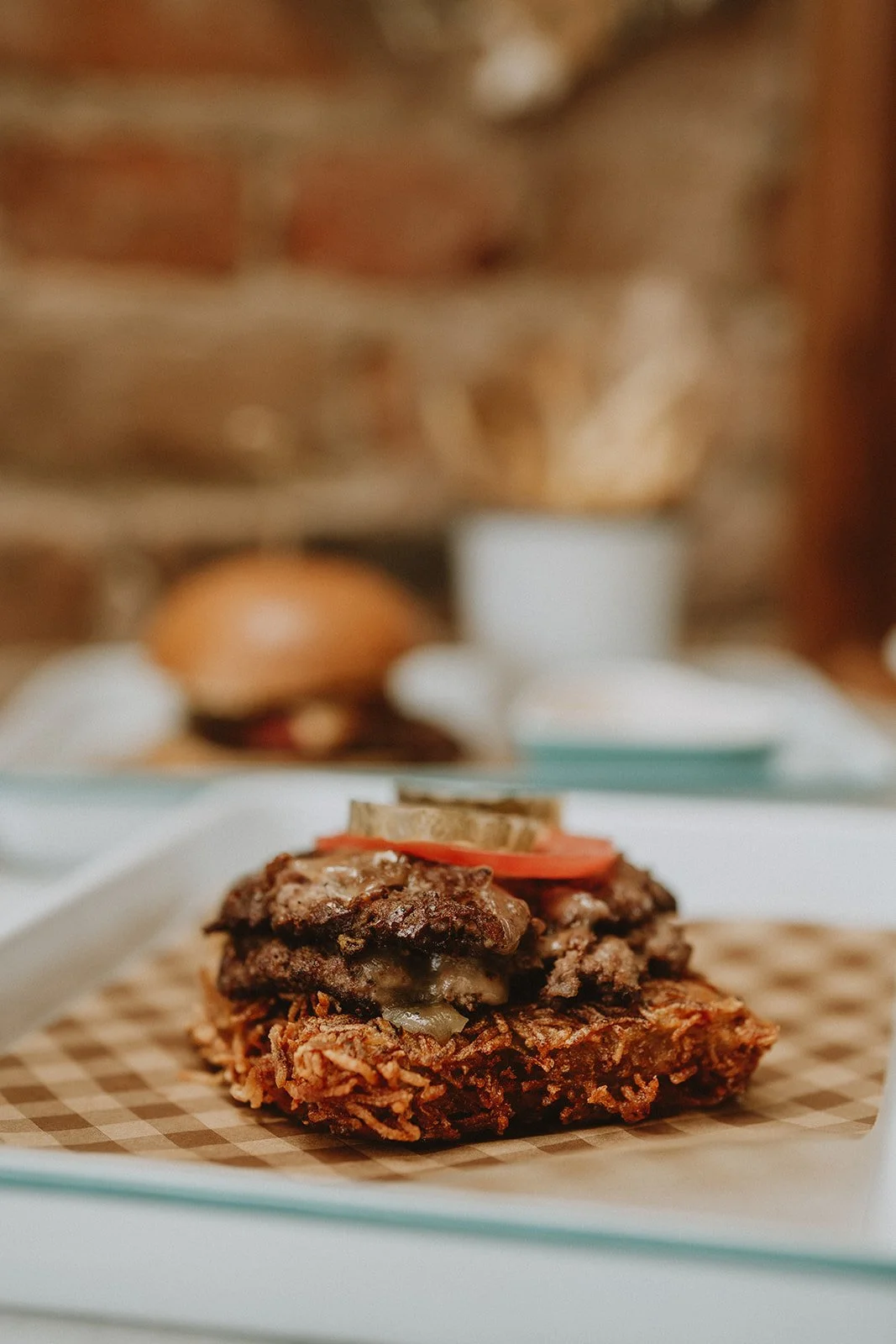 Close-up of a hamburger steak topped with a slice of tomato, pickles, and a slice of onion, on checkered paper, with a burger bun and condiments in the blurred background.