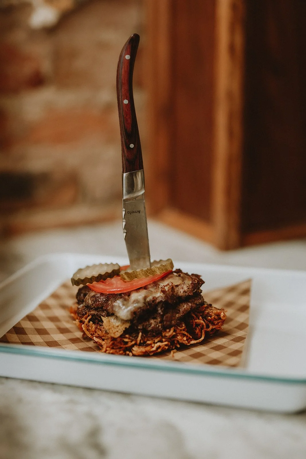 A sliced meatloaf with pickles and tomato slices on top, with a knife inserted into it, on a paper-lined tray.