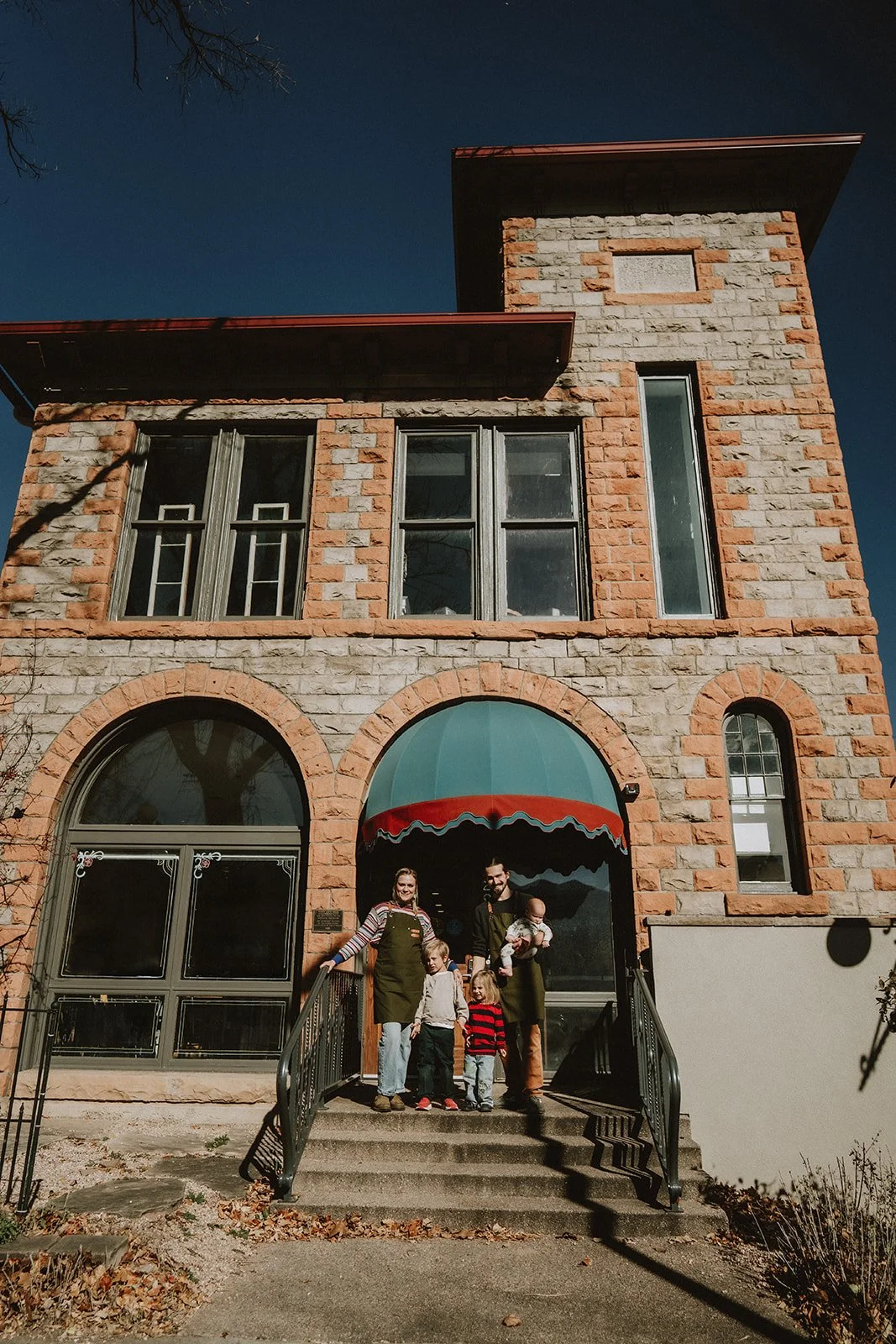 Family of five standing on steps in front of a brick building with arched and rectangular windows, and a green awning. The family includes two adults and three children, one of whom is being held by an adult, under a clear sky.