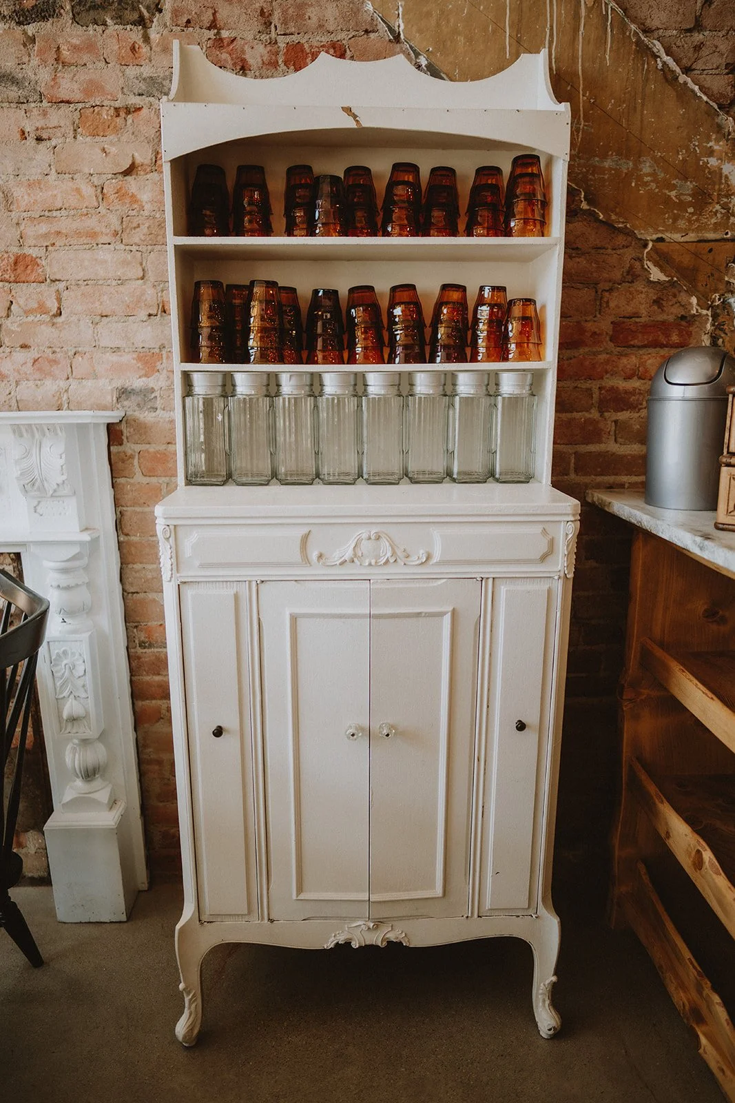 Vintage white wooden cabinet with decorative carved details, holding glass jars and brown glass cups, set against an exposed brick wall.