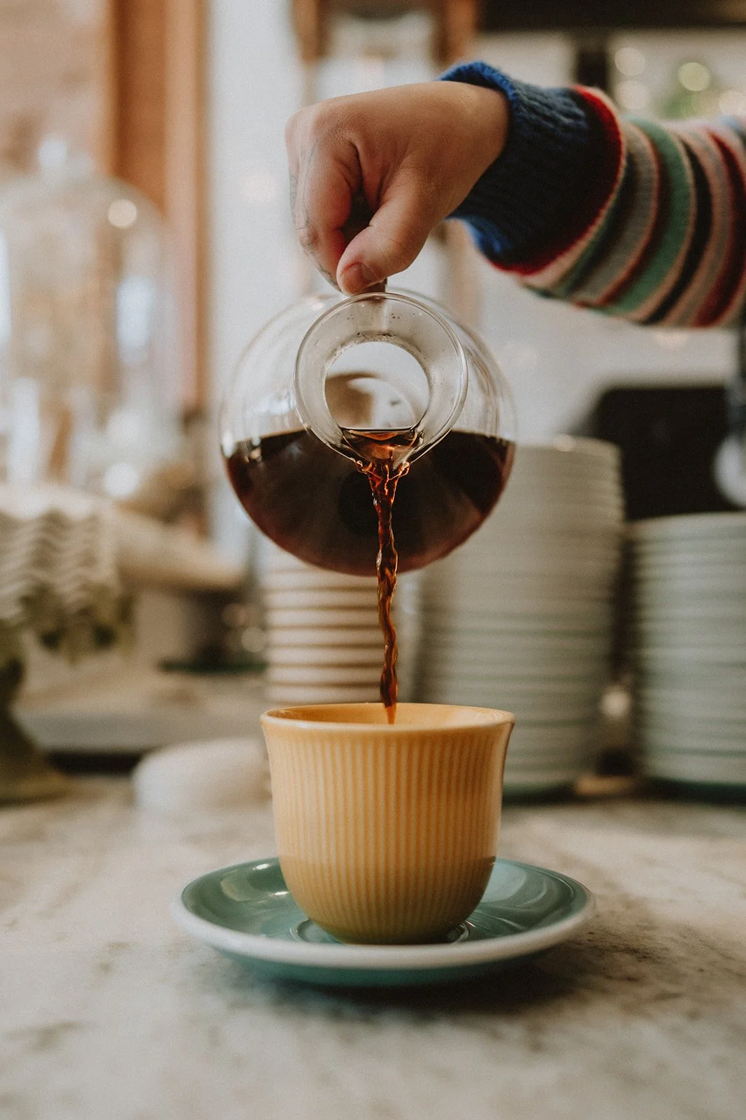 A person wearing a striped sweater is pouring hot coffee from a glass pot into a yellow ceramic cup on a saucer, on a kitchen counter with dishes and kitchen items in the background.