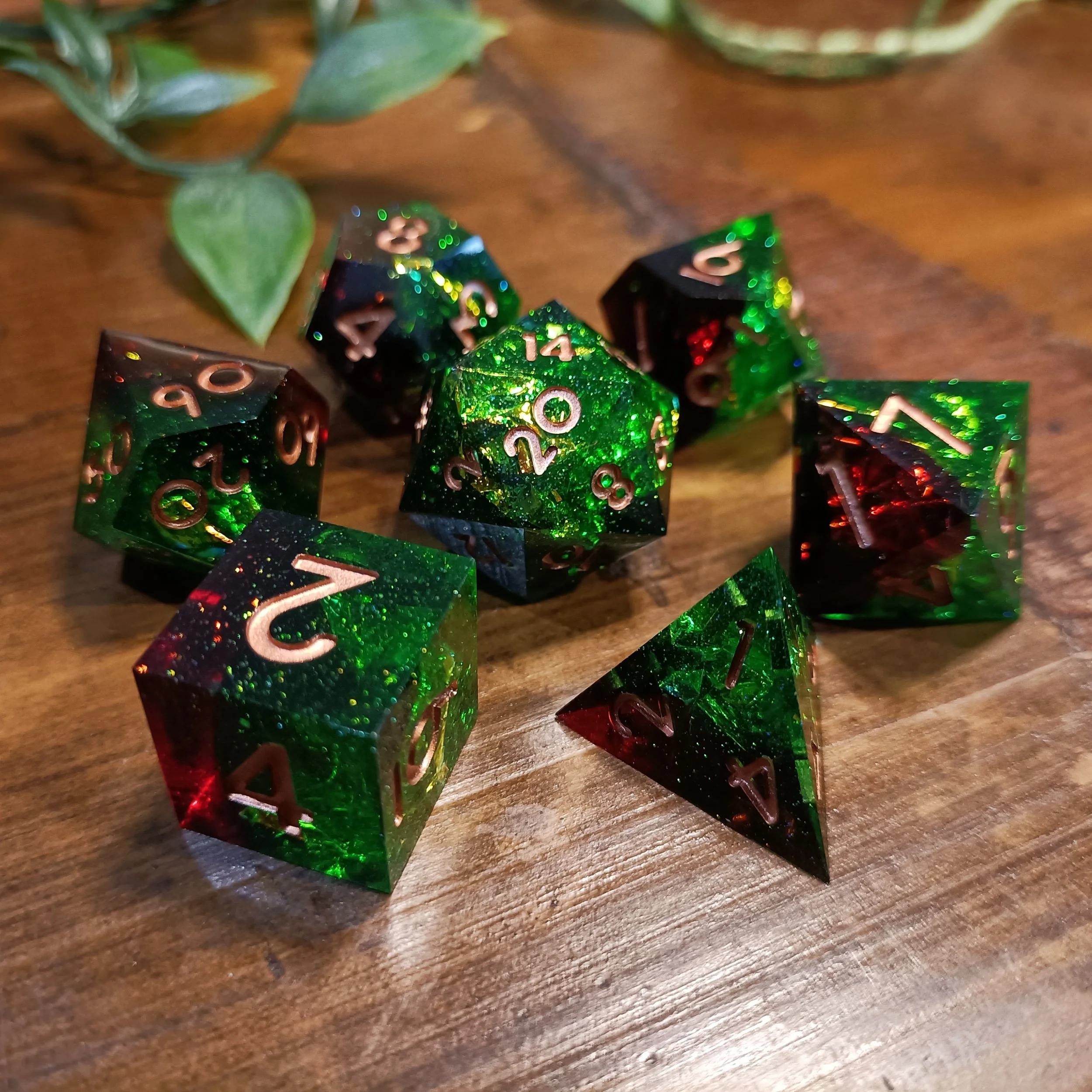 Close-up of a set of half red, half green shiny polyhedral dice on a wooden surface. The dice have metallic bronze numbers and lots of reflective mylar inside.