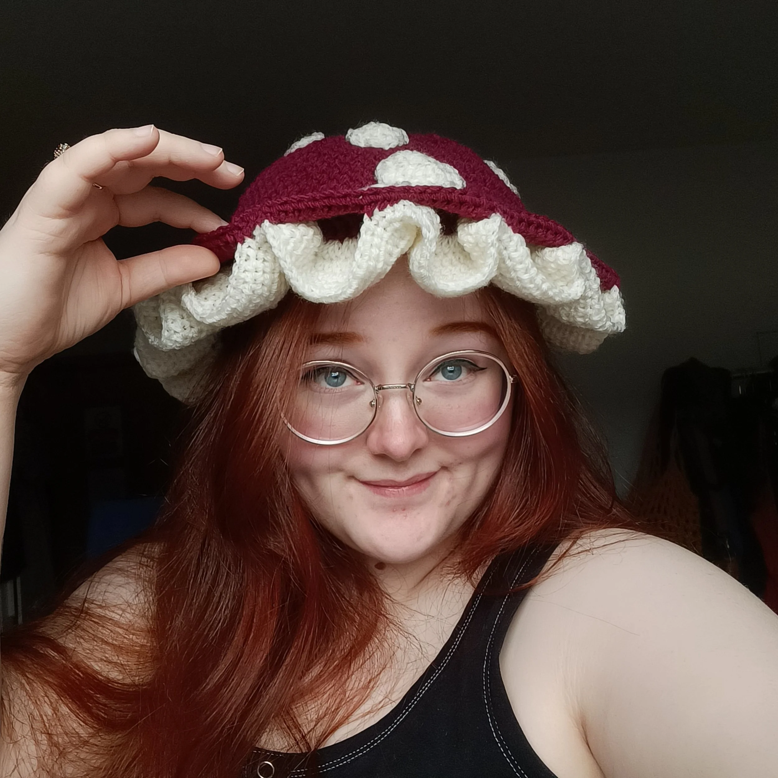 A white woman with red hair wearing a crocheted red mushroom hat with white spots