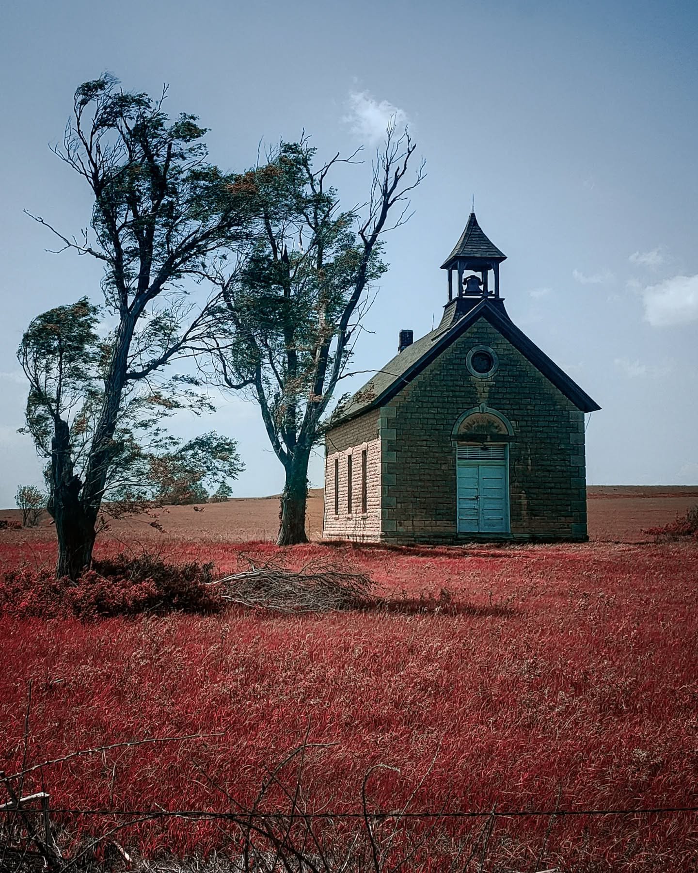 A small stone church with a bell tower is situated in a field with reddish grass. Two trees stand nearby, one mostly bare and the other with sparse leaves, against a blue sky with a few clouds.