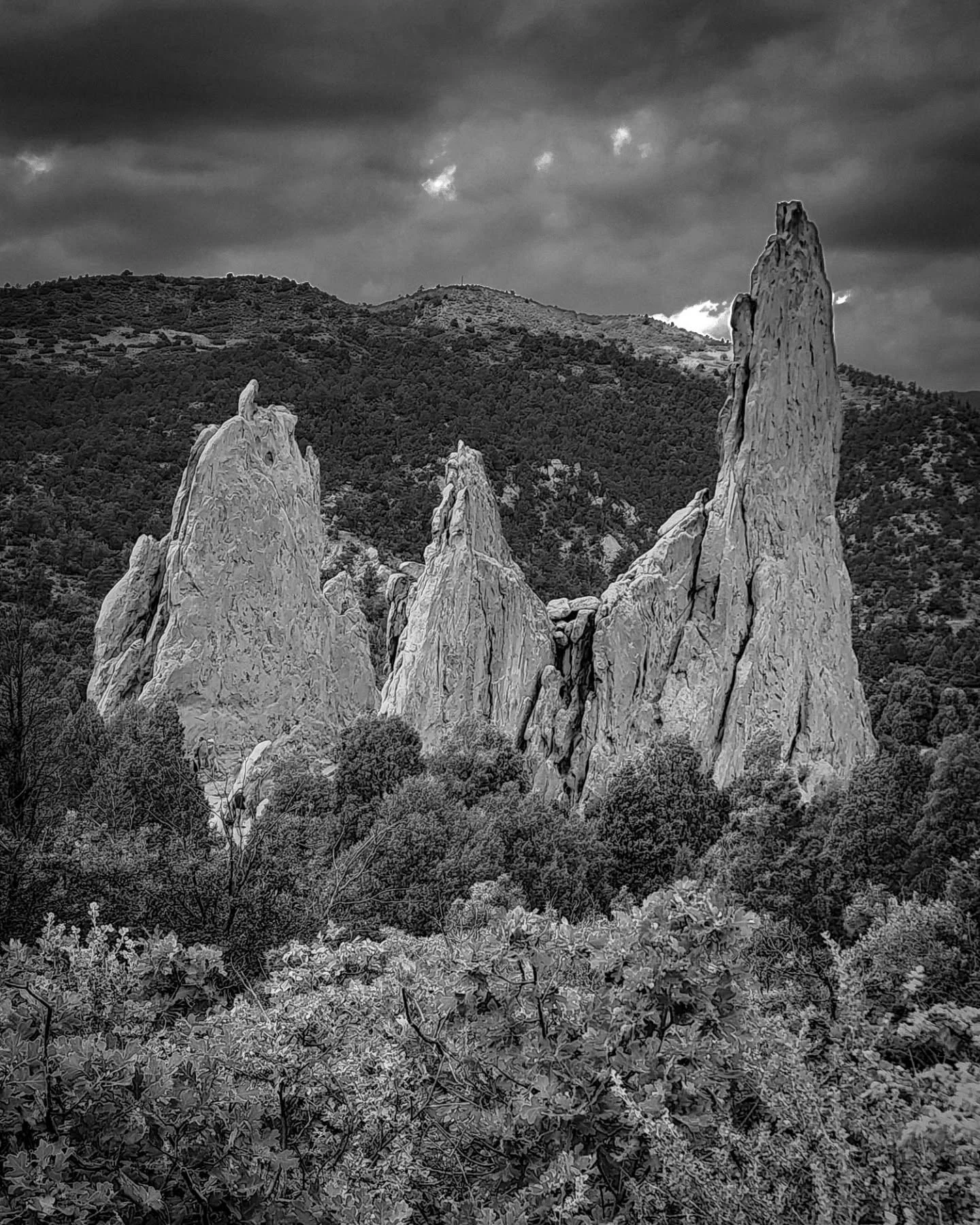 Black and white photograph of three large, jagged rock formations rising from a forested landscape, with a mountain range and cloudy sky in the background.