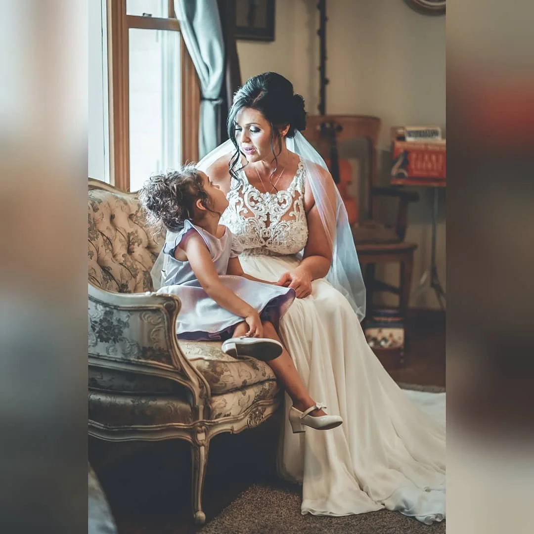 A bride in a wedding dress sitting on a vintage floral sofa with a young girl in a dress and white shoes, engaging in a conversation indoors next to a window with gray curtains.