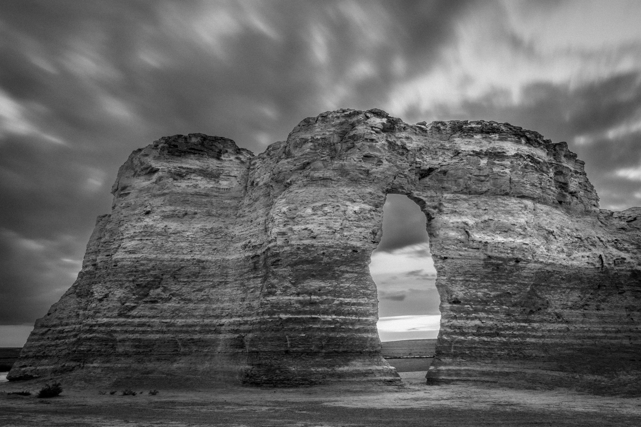 Black and white photo of a natural rock formation with a large arch in the center under a cloudy sky.
