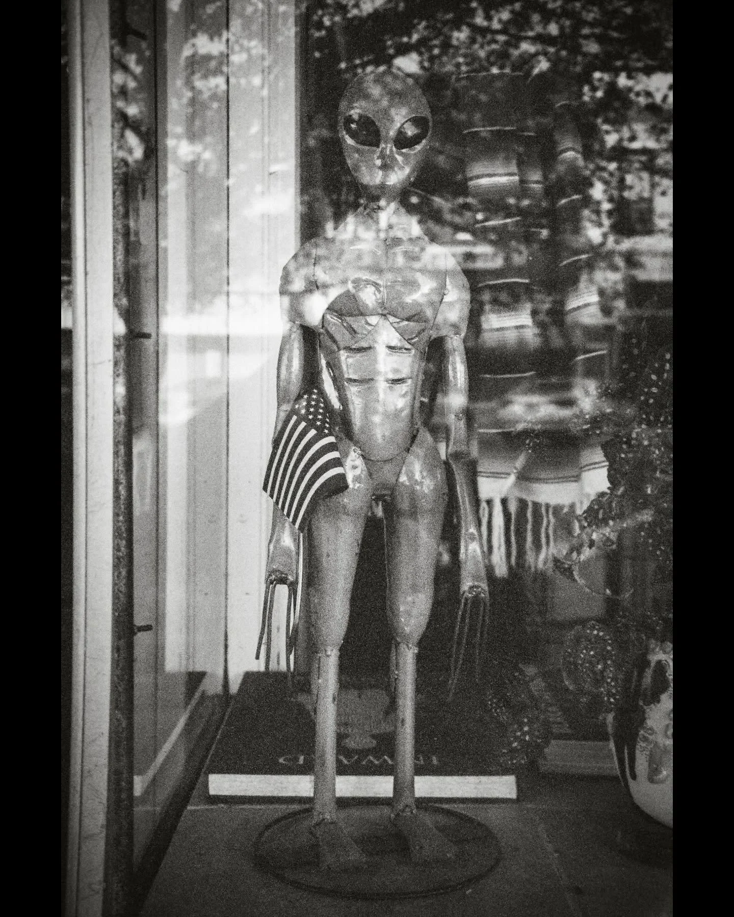 A sculpture of an alien figure with a large head and big eyes, holding a small American flag, displayed behind glass in a window.