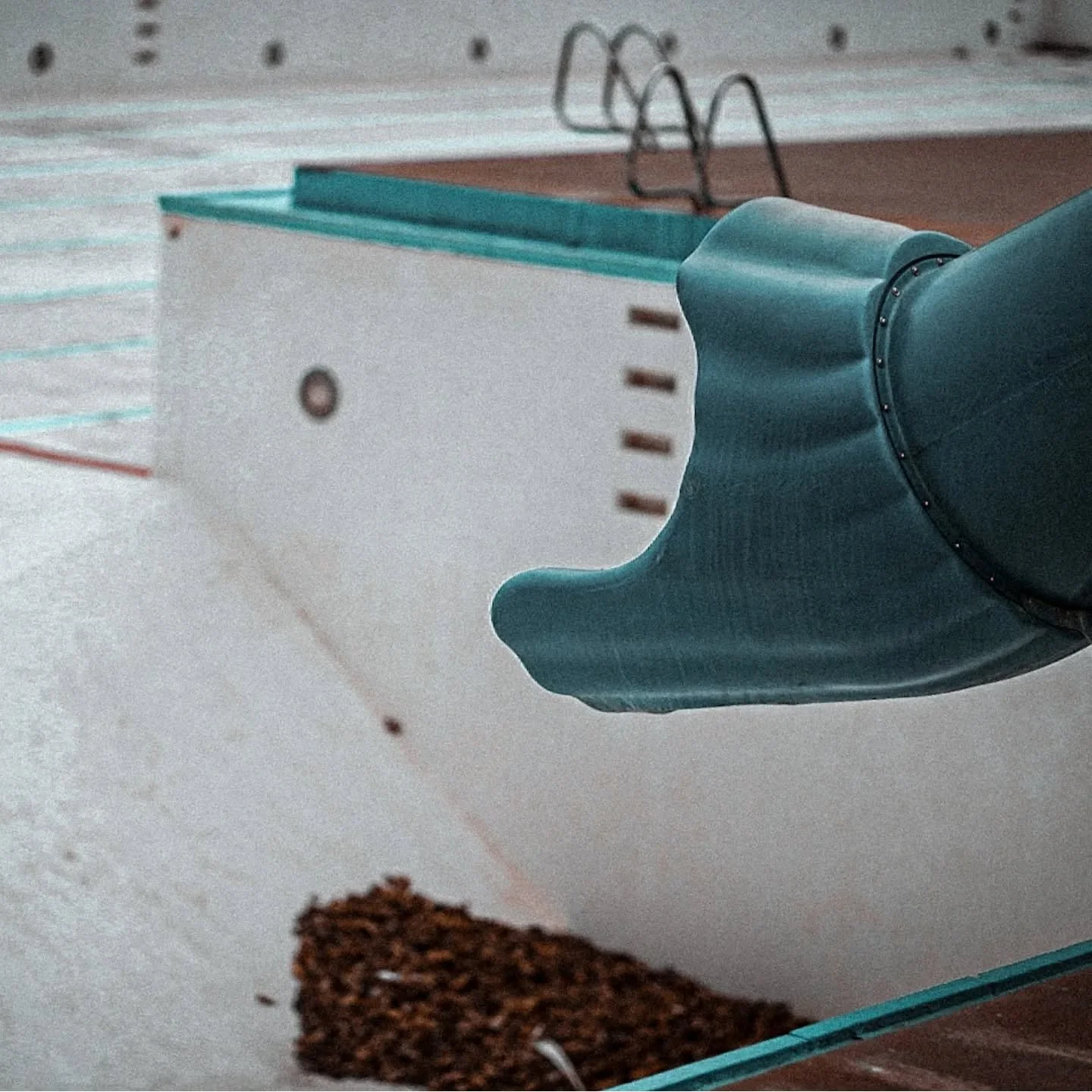 Close-up of a teal-colored slide at a playground with a white and teal structure in the background and brown mulch at the bottom.
