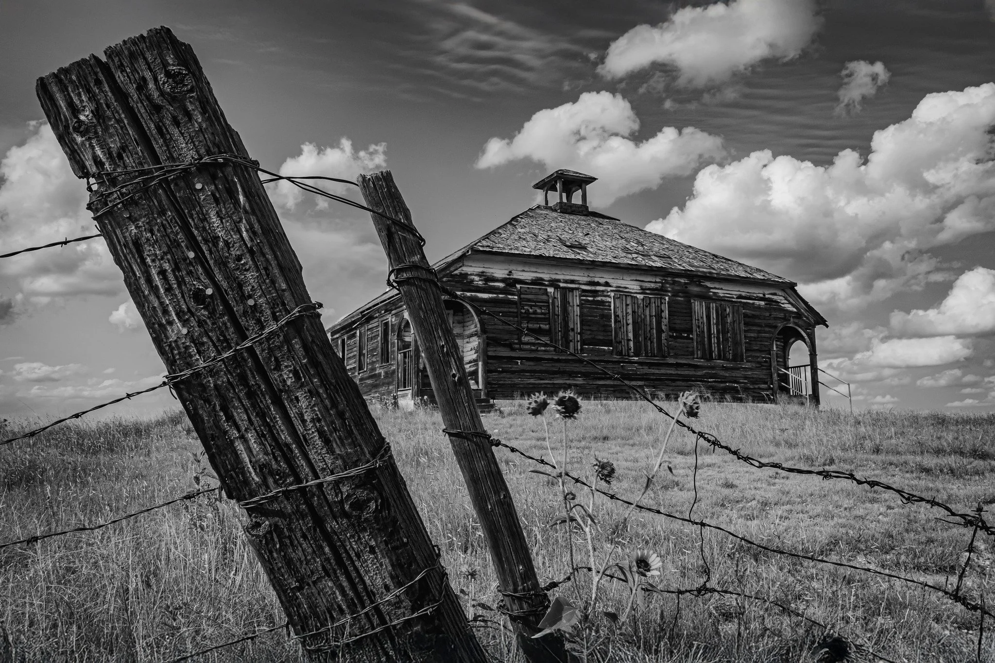 A black and white photo of an old, weathered wooden house in the distance, with a partially broken wooden fence and barbed wire in the foreground. The sky is partly cloudy with large, fluffy clouds.