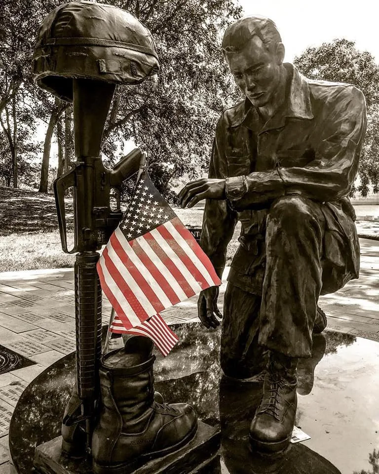 Bronze statue of a soldier kneeling, with a military helmet hanging on a rifle, surrounded by American flags, set outdoors with trees in the background.