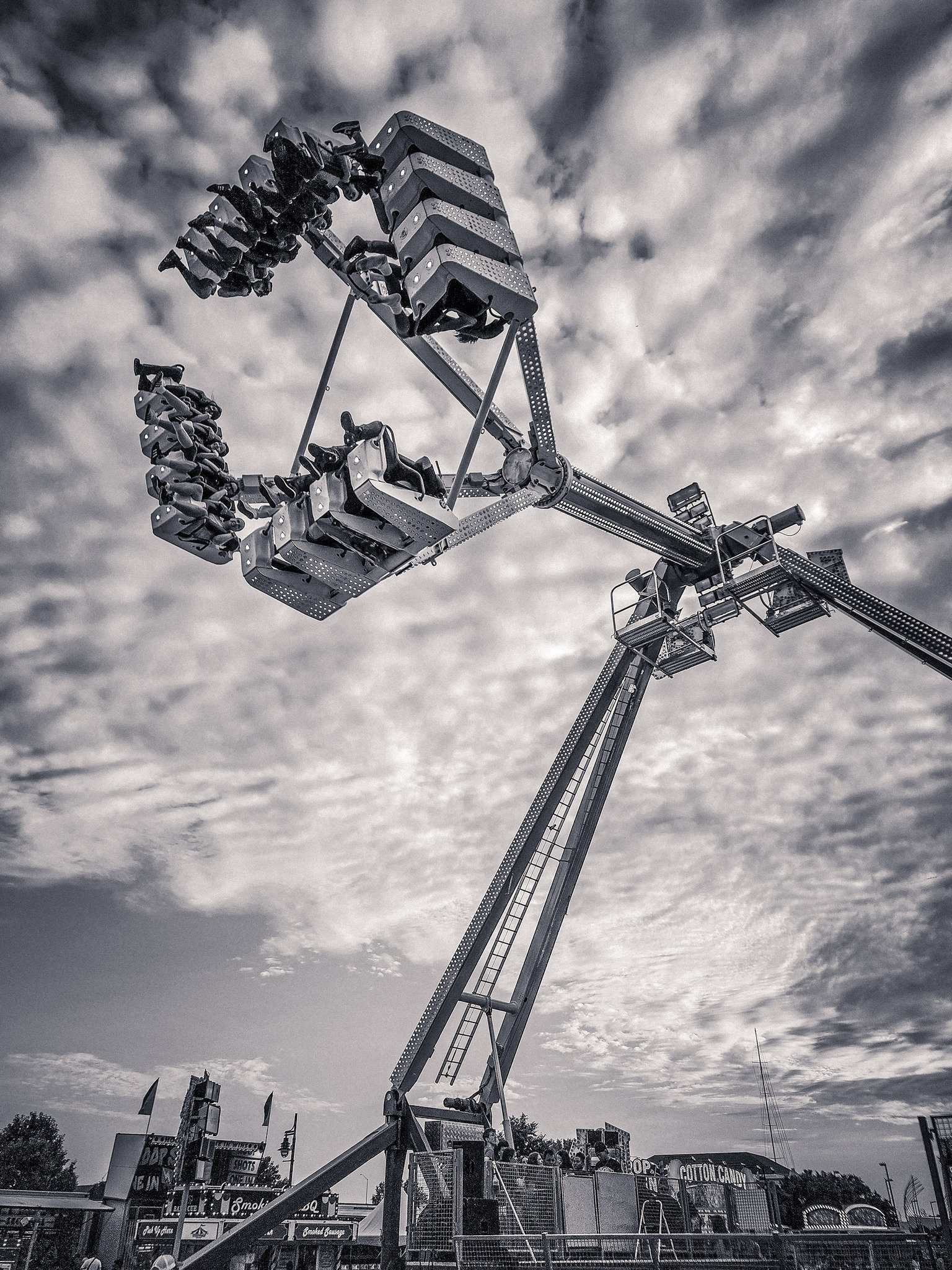 Grayscale photo of a tall amusement park ride with passengers seated in swinging chairs, extended into the sky under a cloudy sky.