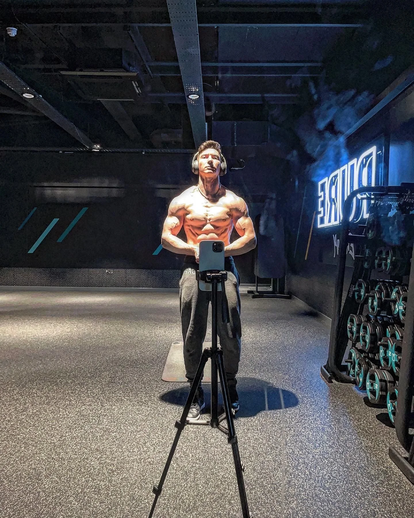 A muscular young man wearing a headset and black pants takes a selfie in a gym mirror, flexing his muscles with gym equipment and a neon sign visible in the background.