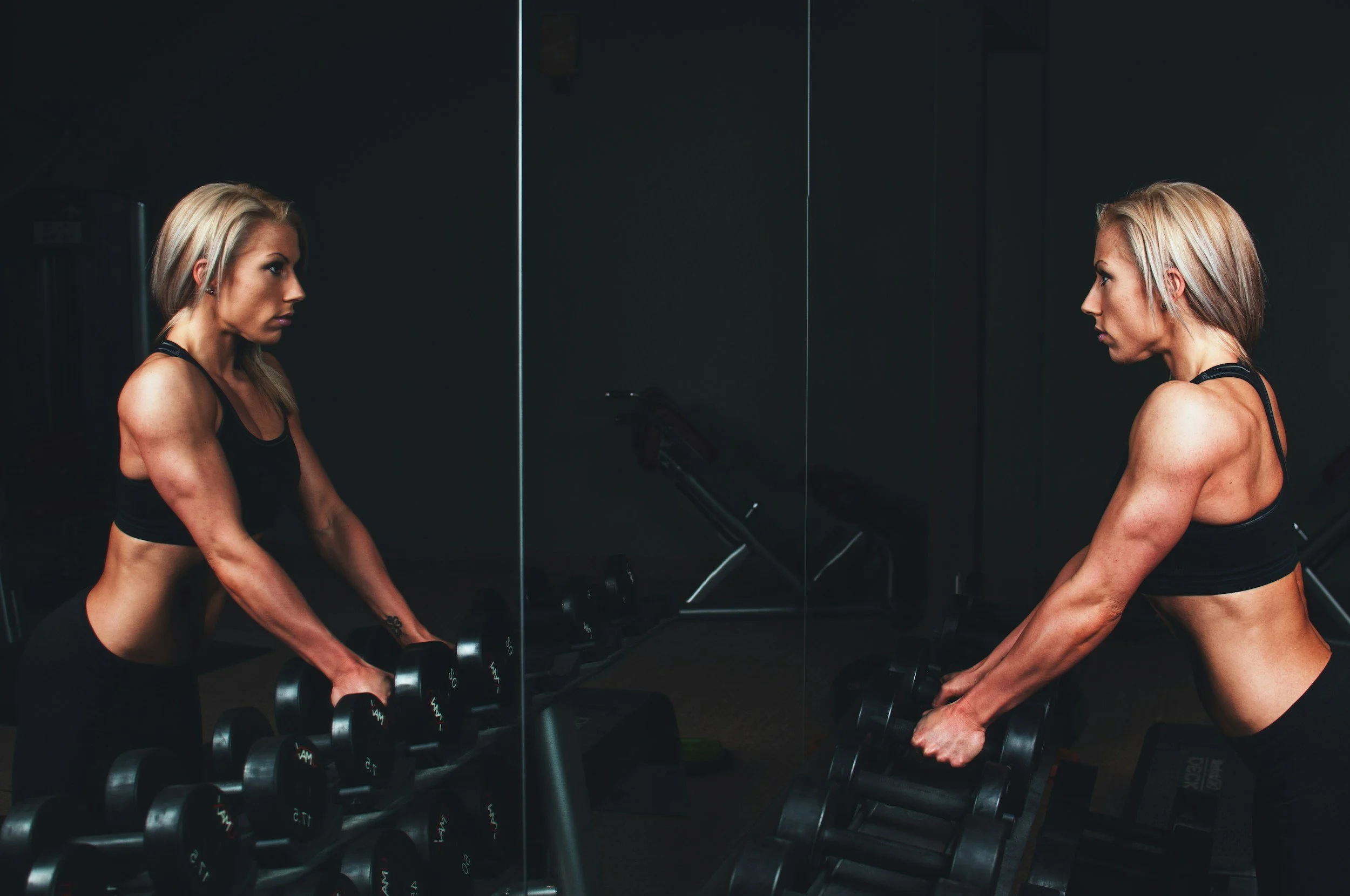 A fit woman with blond hair doing a workout with dumbbells in a gym, standing in front of a mirror