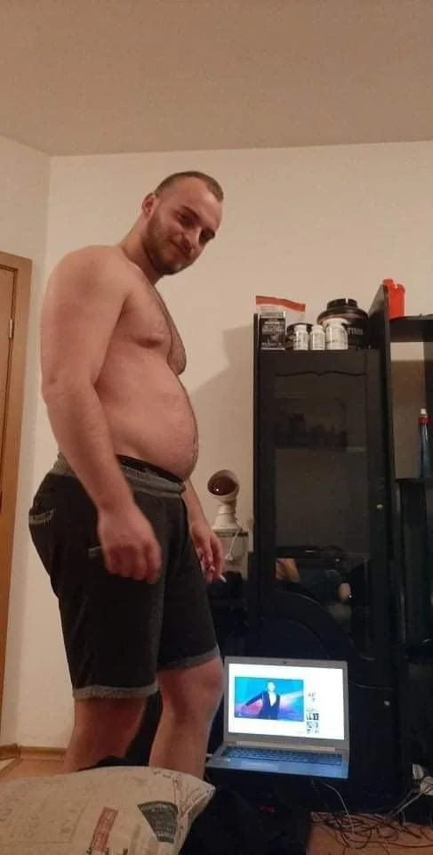 A shirtless man with a beard standing in a room with a laptop displaying a news anchor, surrounded by various objects on shelves.