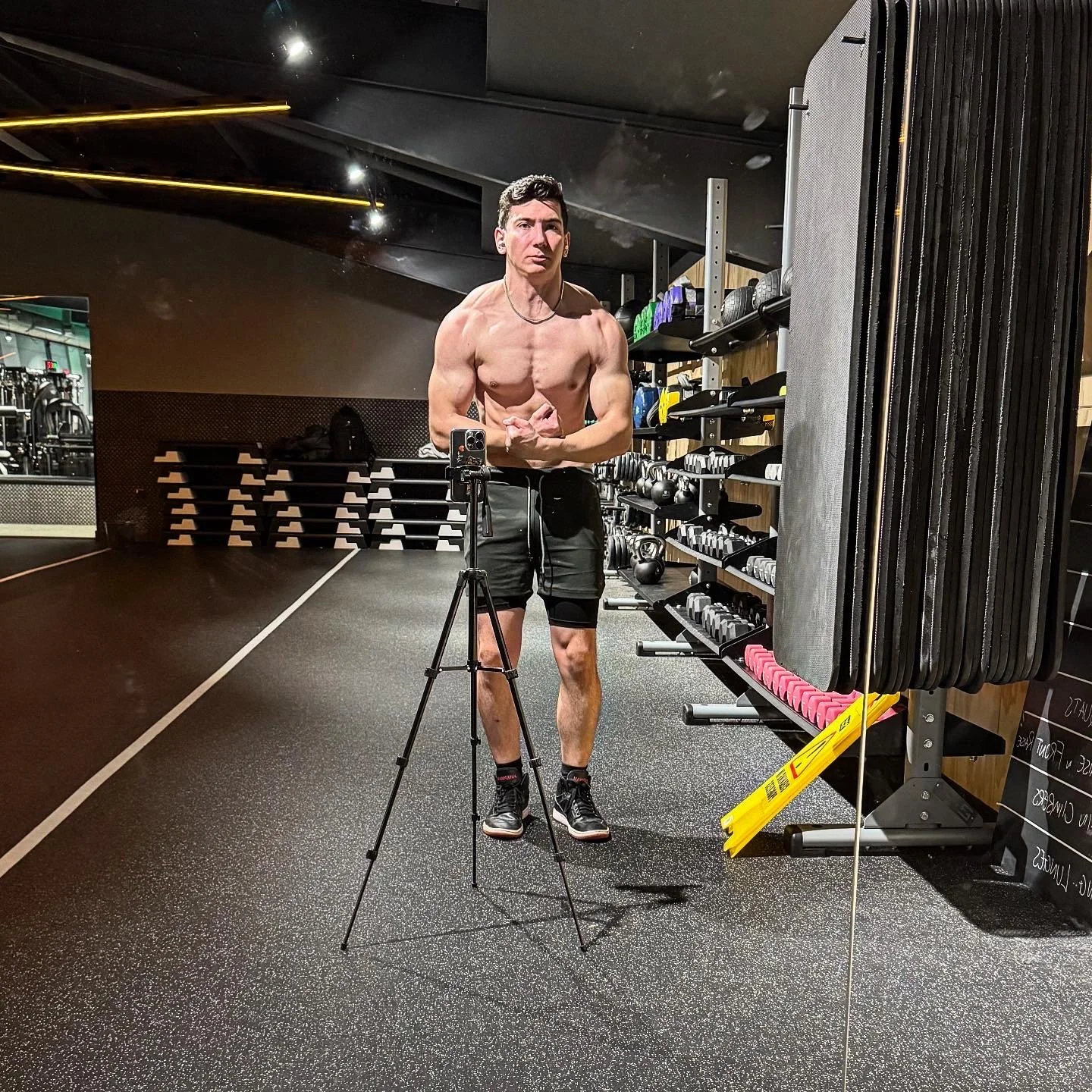 A shirtless young man with muscular build taking a selfie in a gym mirror, standing on black rubber flooring, with shelves of kettlebells and dumbbells behind him and black fitness mats stacked to the right.