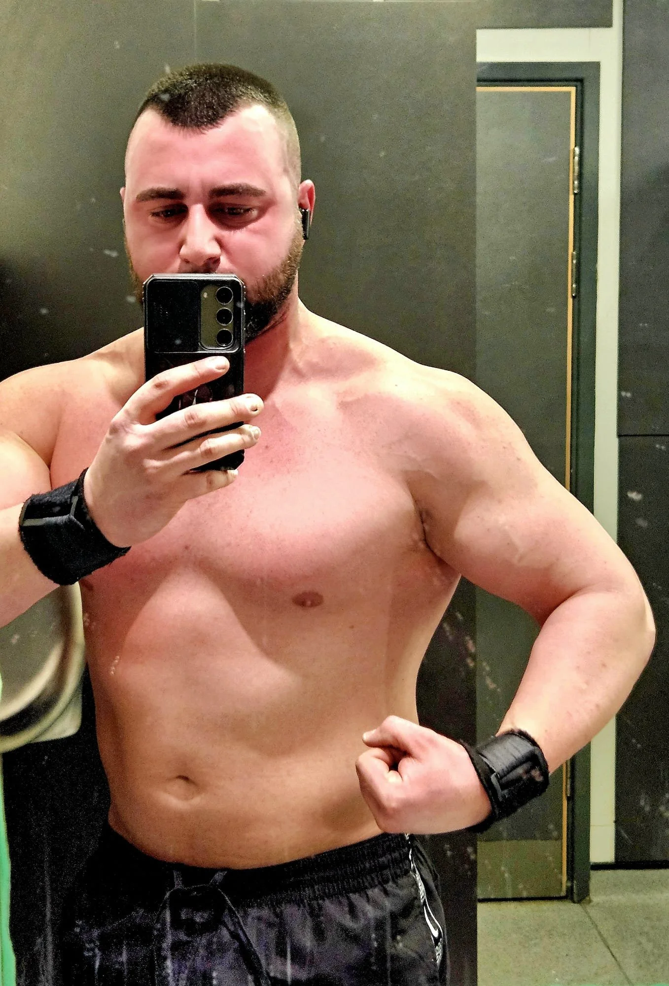 A shirtless man taking a mirror selfie in a gym locker room, flexing his arm with a serious expression.