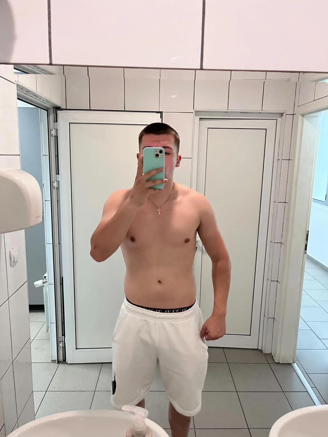A young man taking a mirror selfie in a bathroom, shirtless, wearing white shorts with black accents, and a cross necklace.