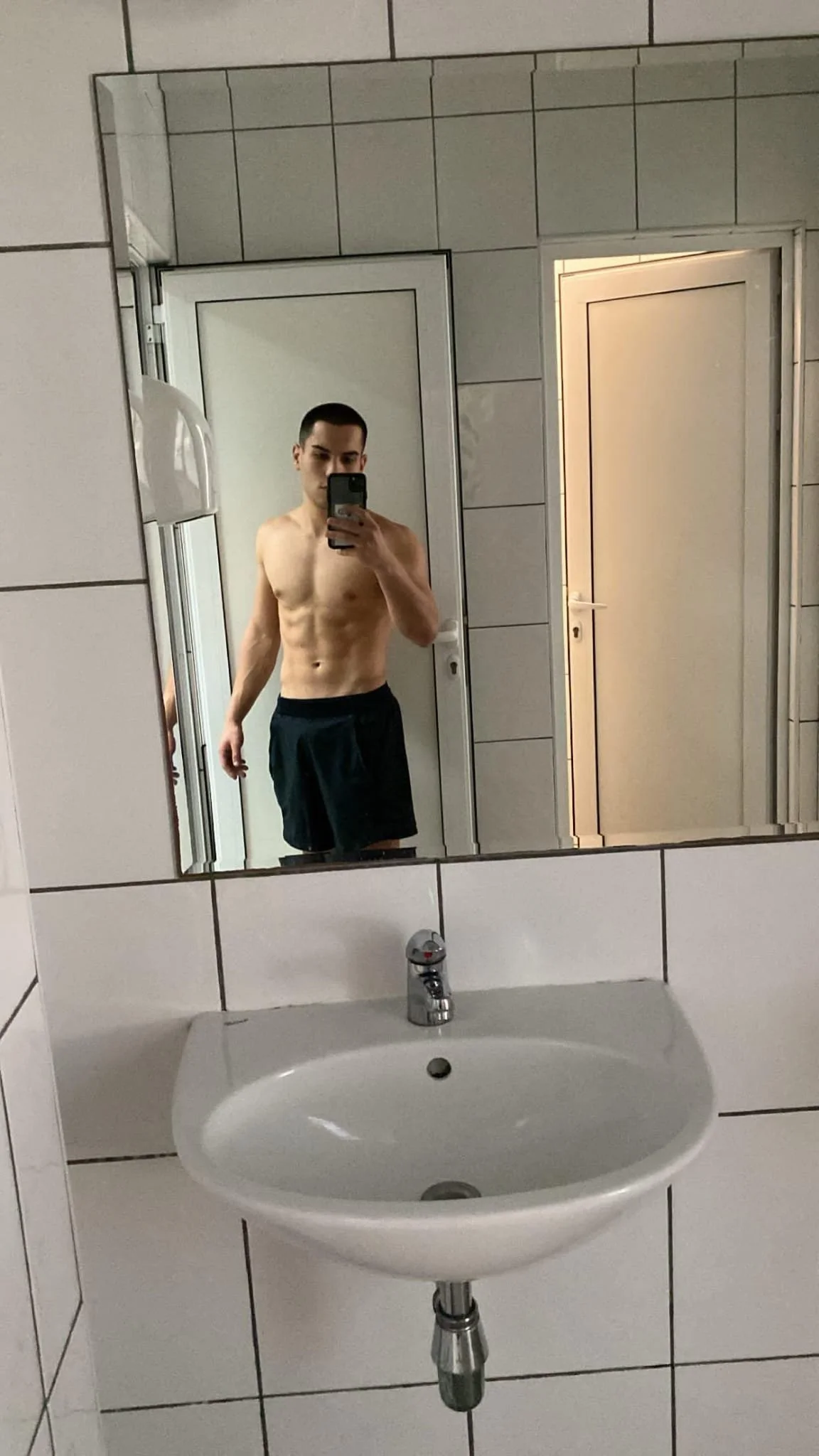 A shirtless young man taking a selfie in a bathroom mirror, wearing black shorts.