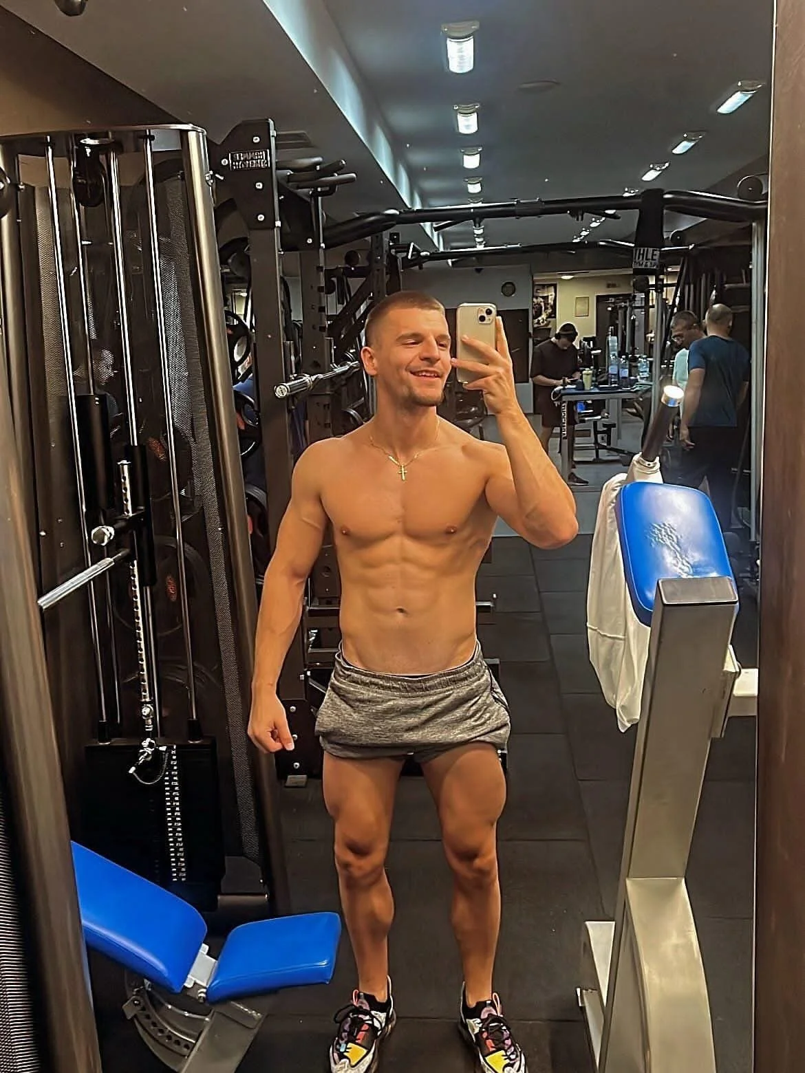 A shirtless man taking a selfie in a gym mirror, wearing shorts and sneakers, with gym equipment and other people working out in the background.