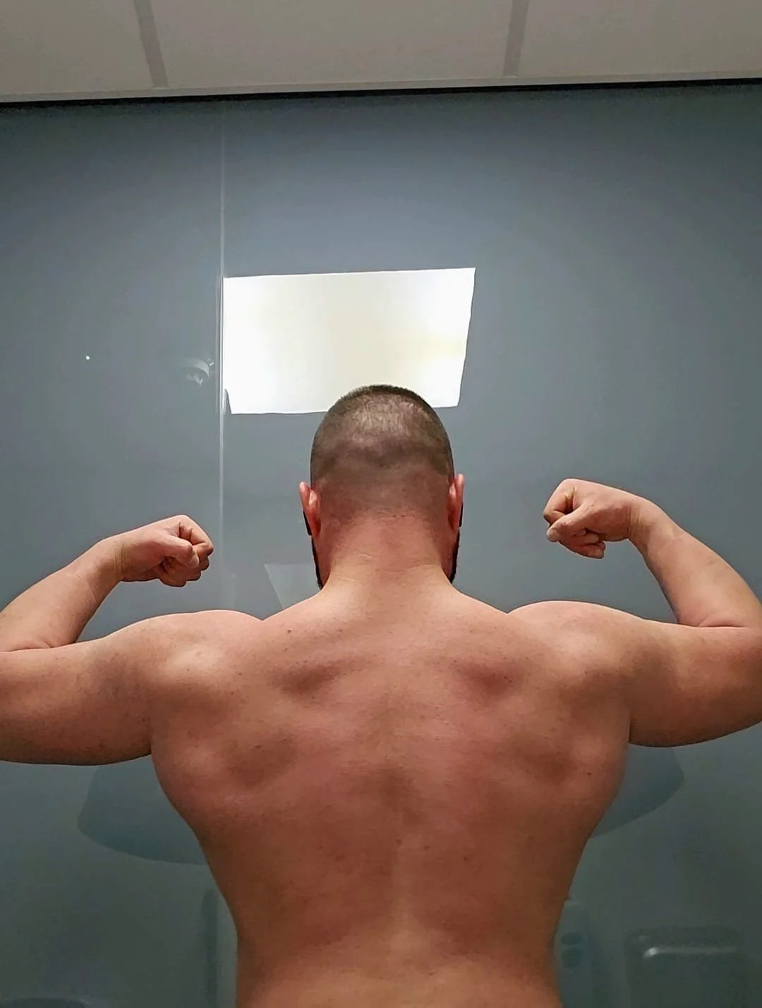 Back view of a muscular man flexing his biceps in front of a mirror.