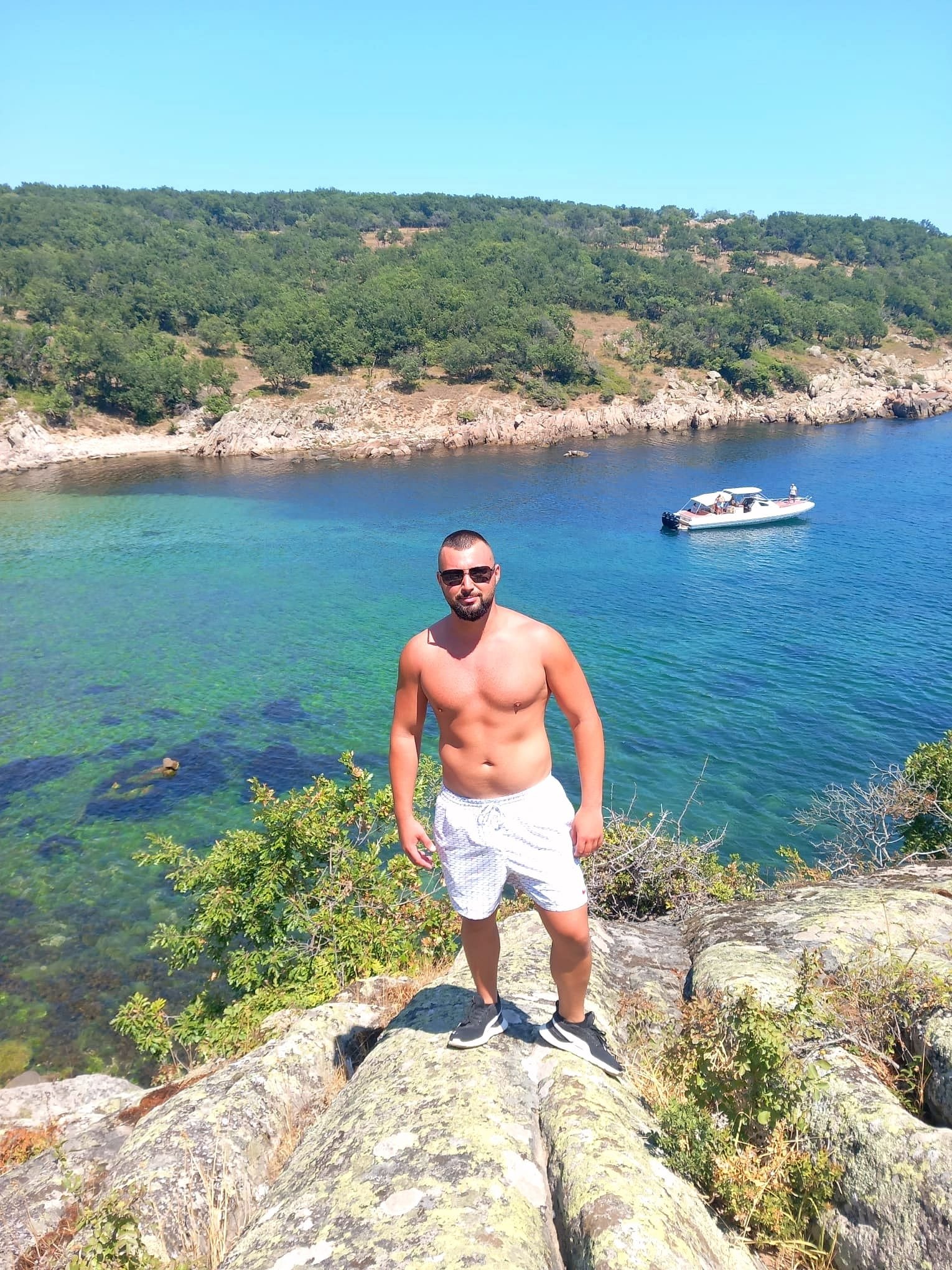 A shirtless man wearing sunglasses, white shorts, and sneakers standing on rocks near a body of water with a boat in the background and a green, forested hillside under a clear blue sky.