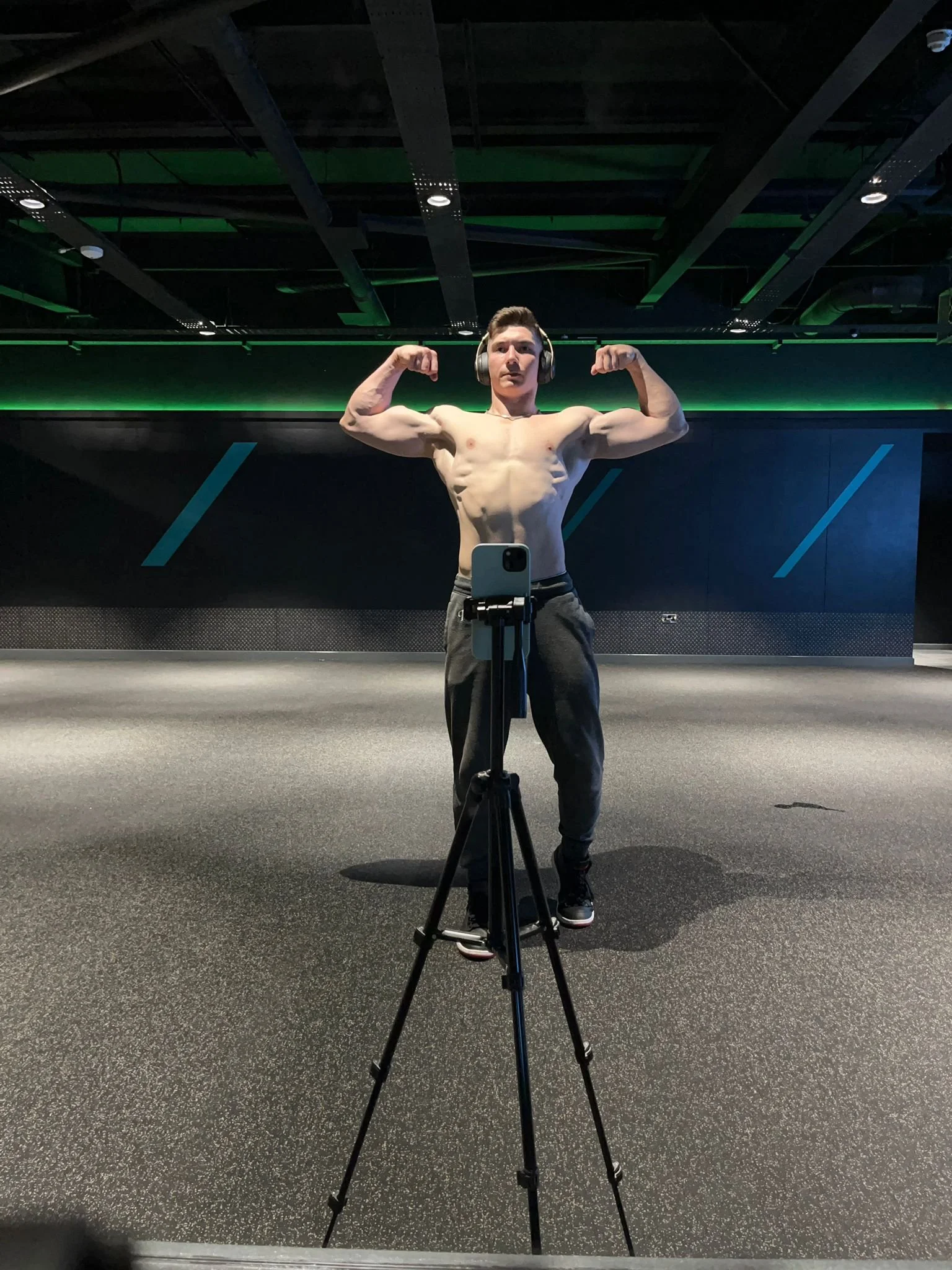 A shirtless young man wearing headphones is flexing his biceps in front of a mirror at a gym, taking a photo with a smartphone on a tripod.