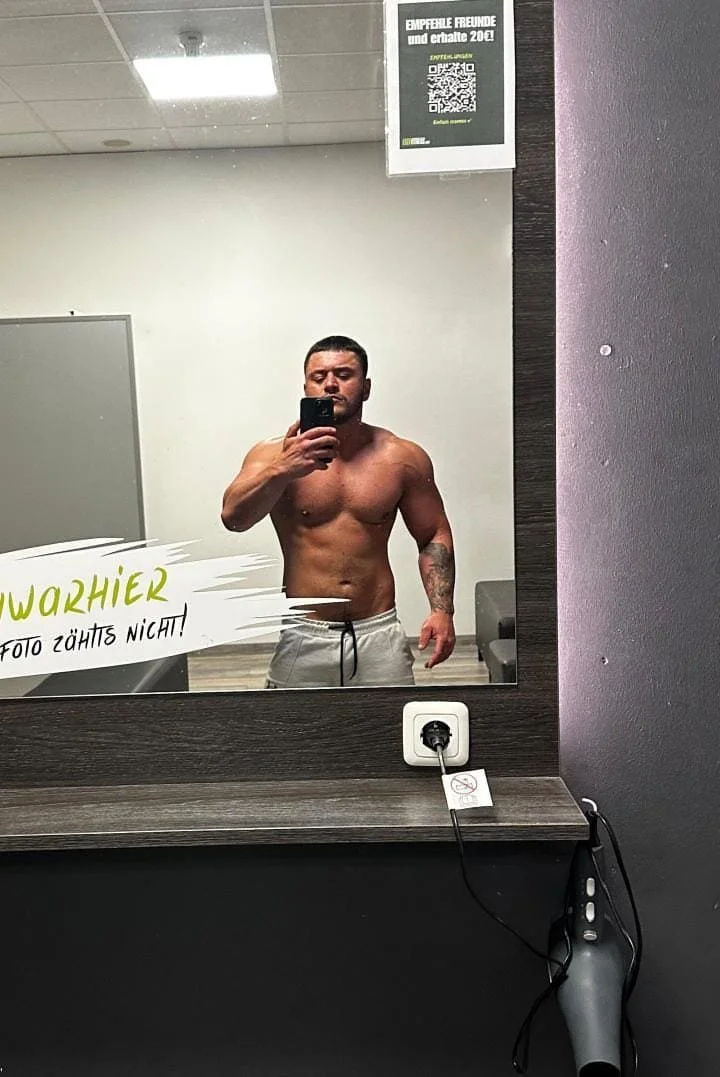 A shirtless man taking a selfie in a mirror at a gym or locker room.