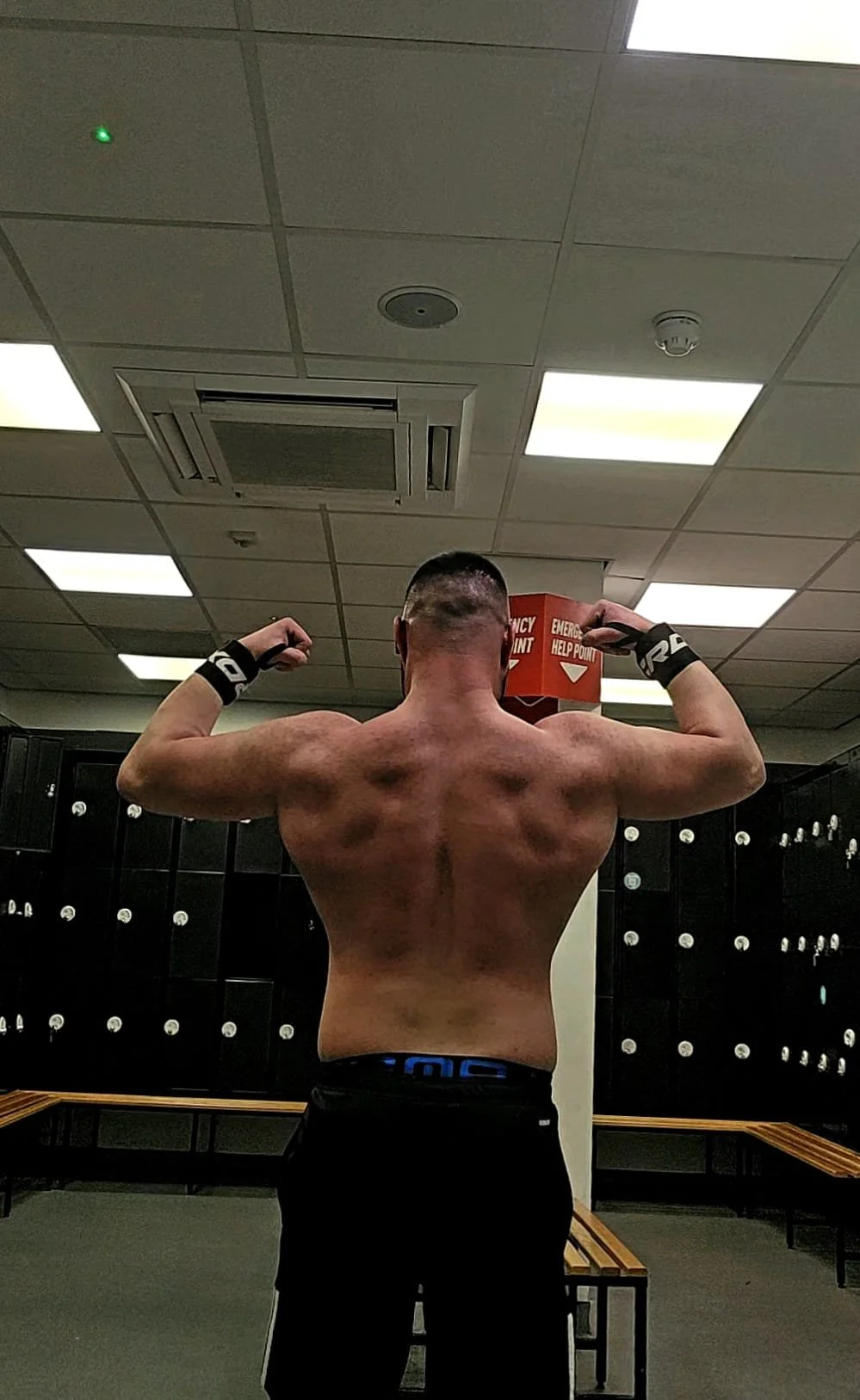 A shirtless man with muscular physique and short hair flexing his biceps in a locker room, with lockers and benches in the background.