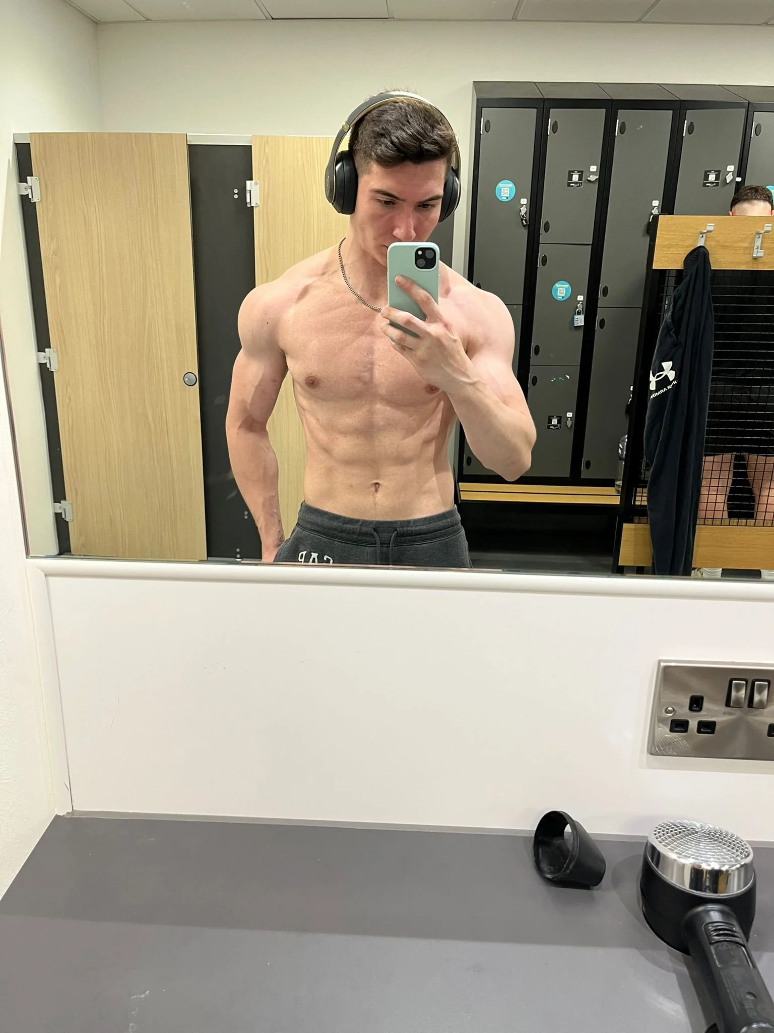 A shirtless young man wearing headphones taking a mirror selfie in a gym locker room, with black lockers and wooden benches in the background.
