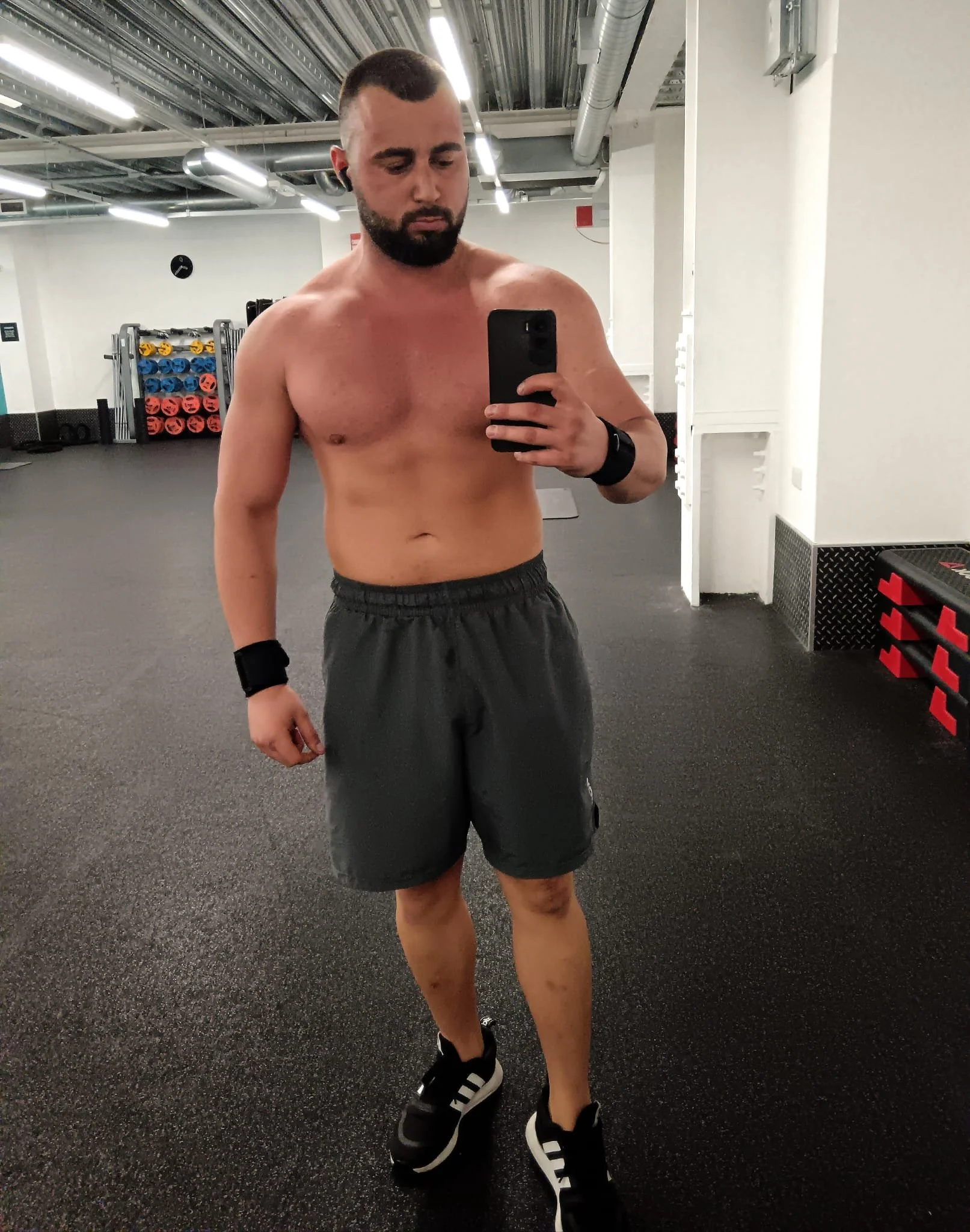 A shirtless man taking a selfie in a gym mirror, wearing black shorts, black sneakers with white stripes, black wristbands, and wireless earbuds, with gym equipment in the background.