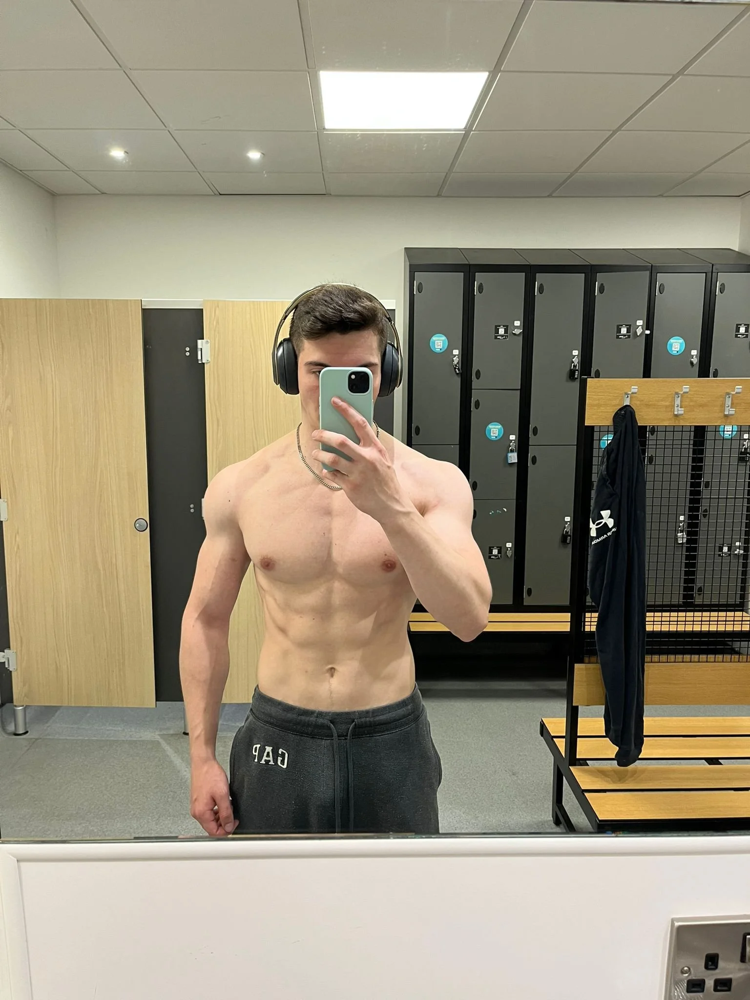 A shirtless young man in a gym locker room taking a mirror selfie with his phone, wearing headphones and gray sweatpants.
