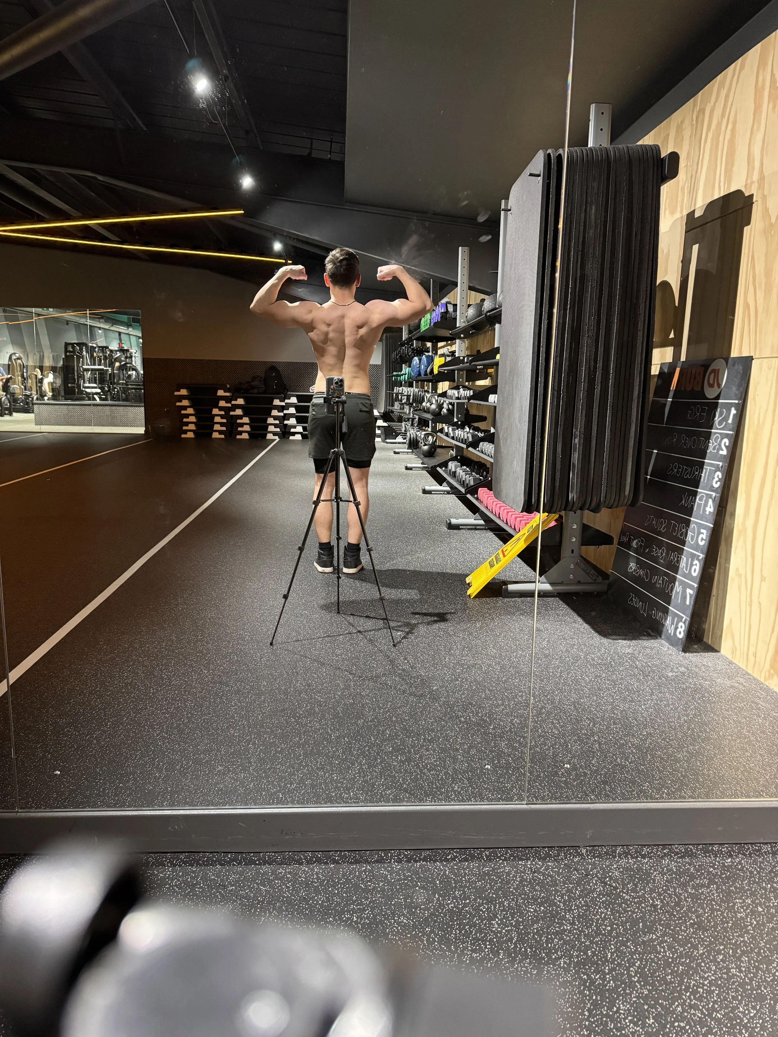 A shirtless man taking a photo of his back muscles in a gym mirror, with gym equipment and a blackboard in the background.
