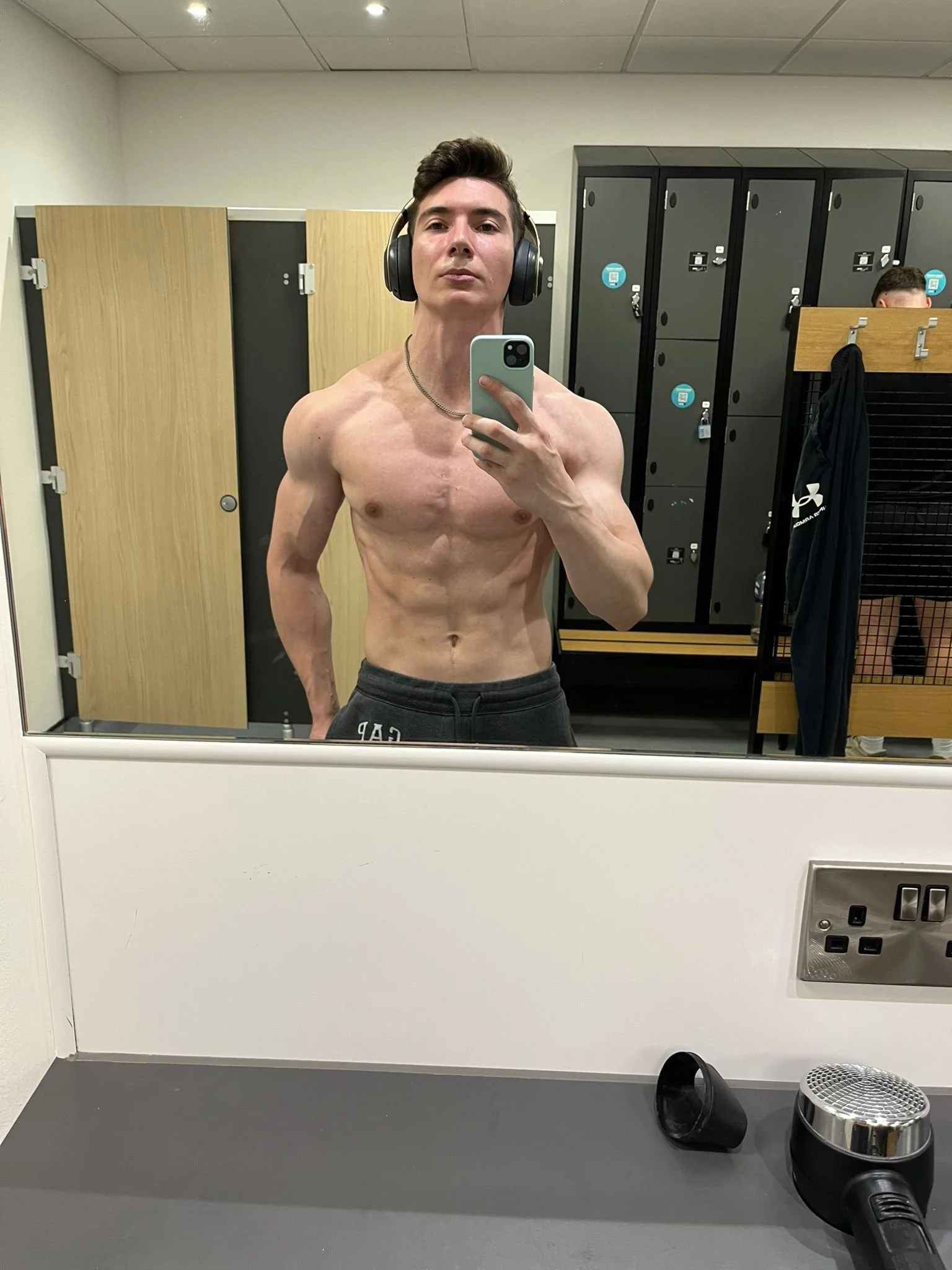 A shirtless young man with headphones taking a selfie in a gym locker room, with lockers and another person in the background.