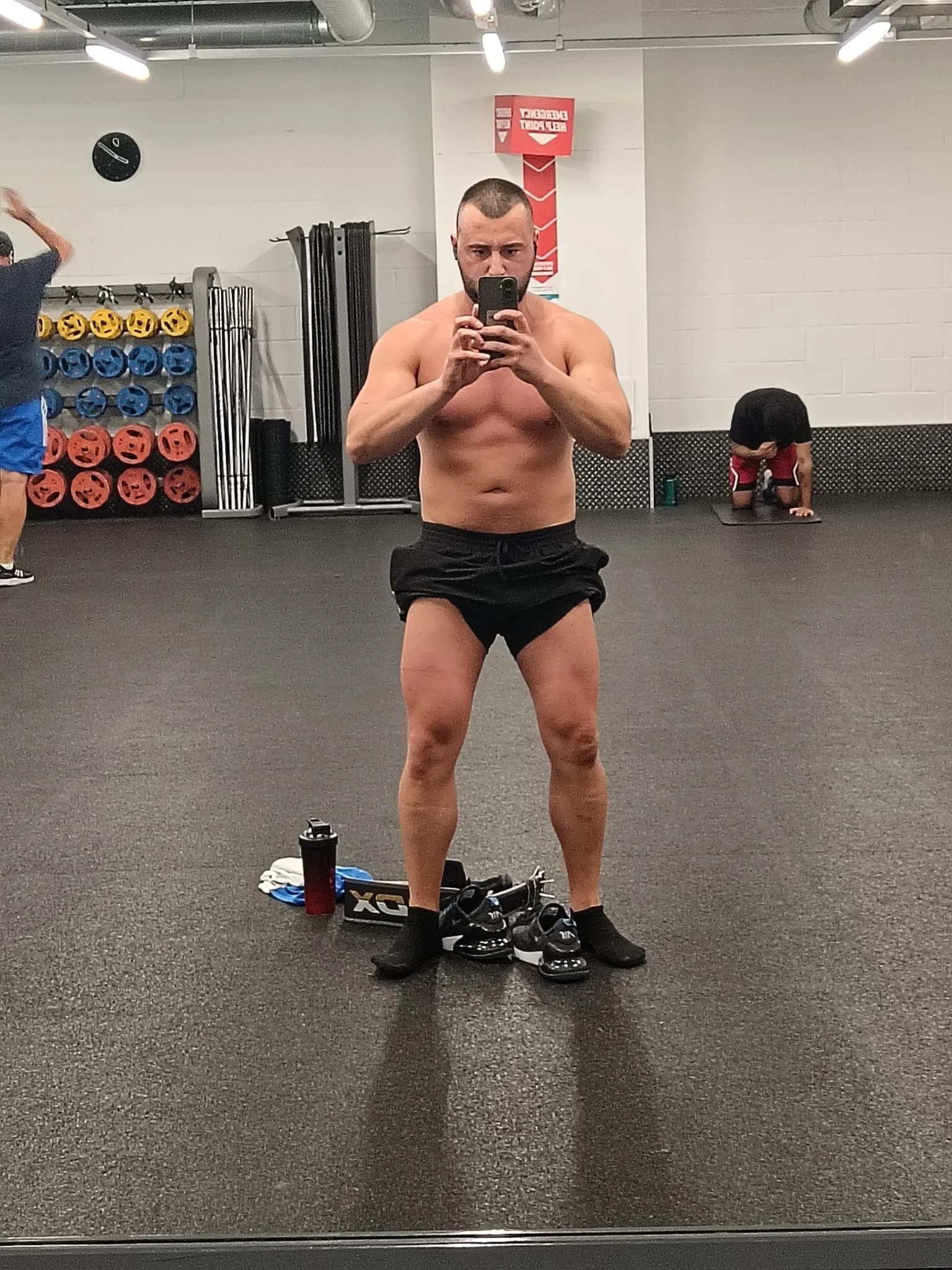A shirtless man taking a selfie in a gym mirror, wearing black shorts and black socks, with workout shoes and fitness gear on the floor in front of him. Background includes gym equipment and other people exercising.