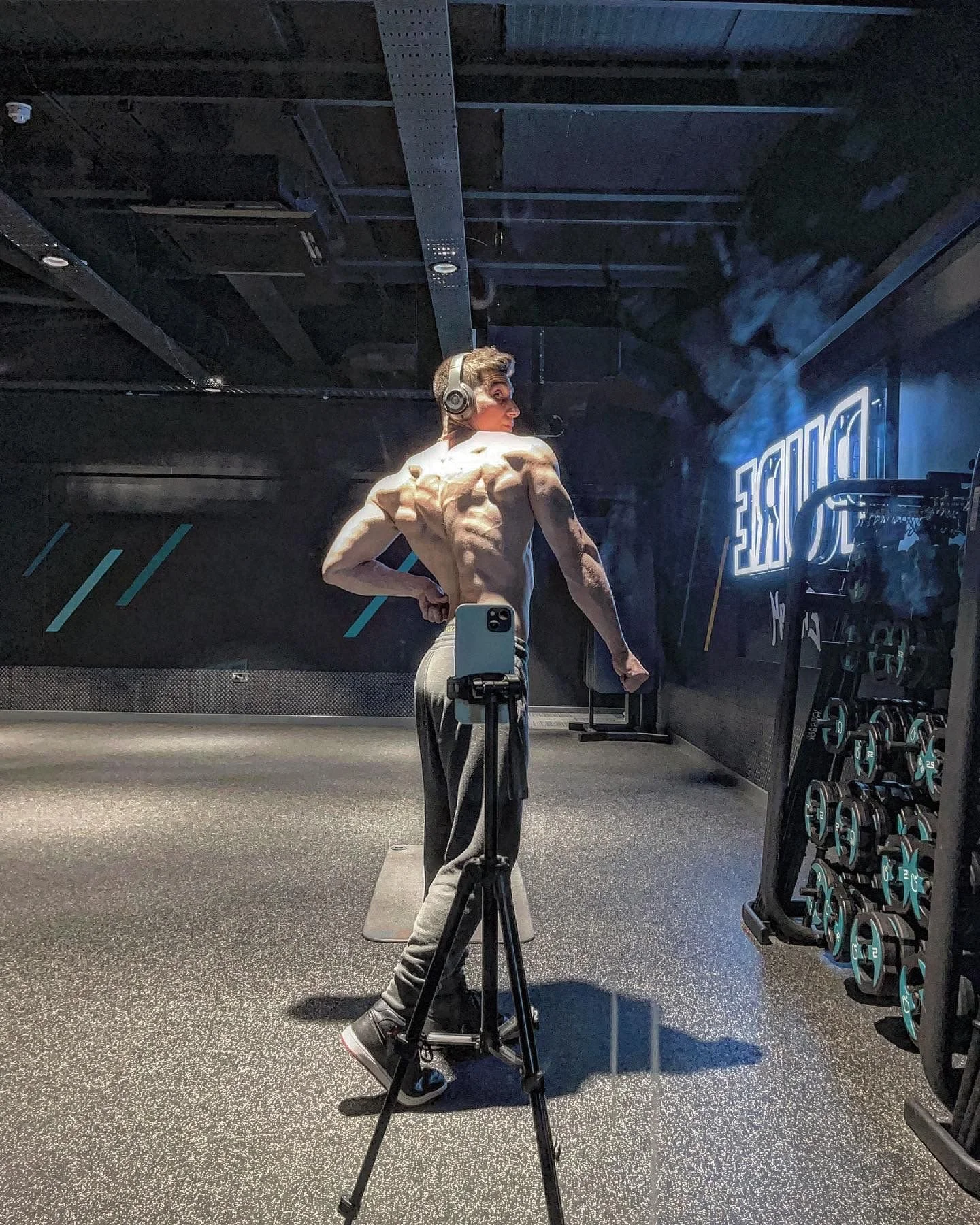 A shirtless man with tattooed arms and lean muscles taking a mirror selfie in a gym, using a tripod phone holder. The man is standing in front of a dark wall with blue diagonal lines and a neon sign, and his reflection is visible in the mirror. He is wearing gray sweatpants and headphones.