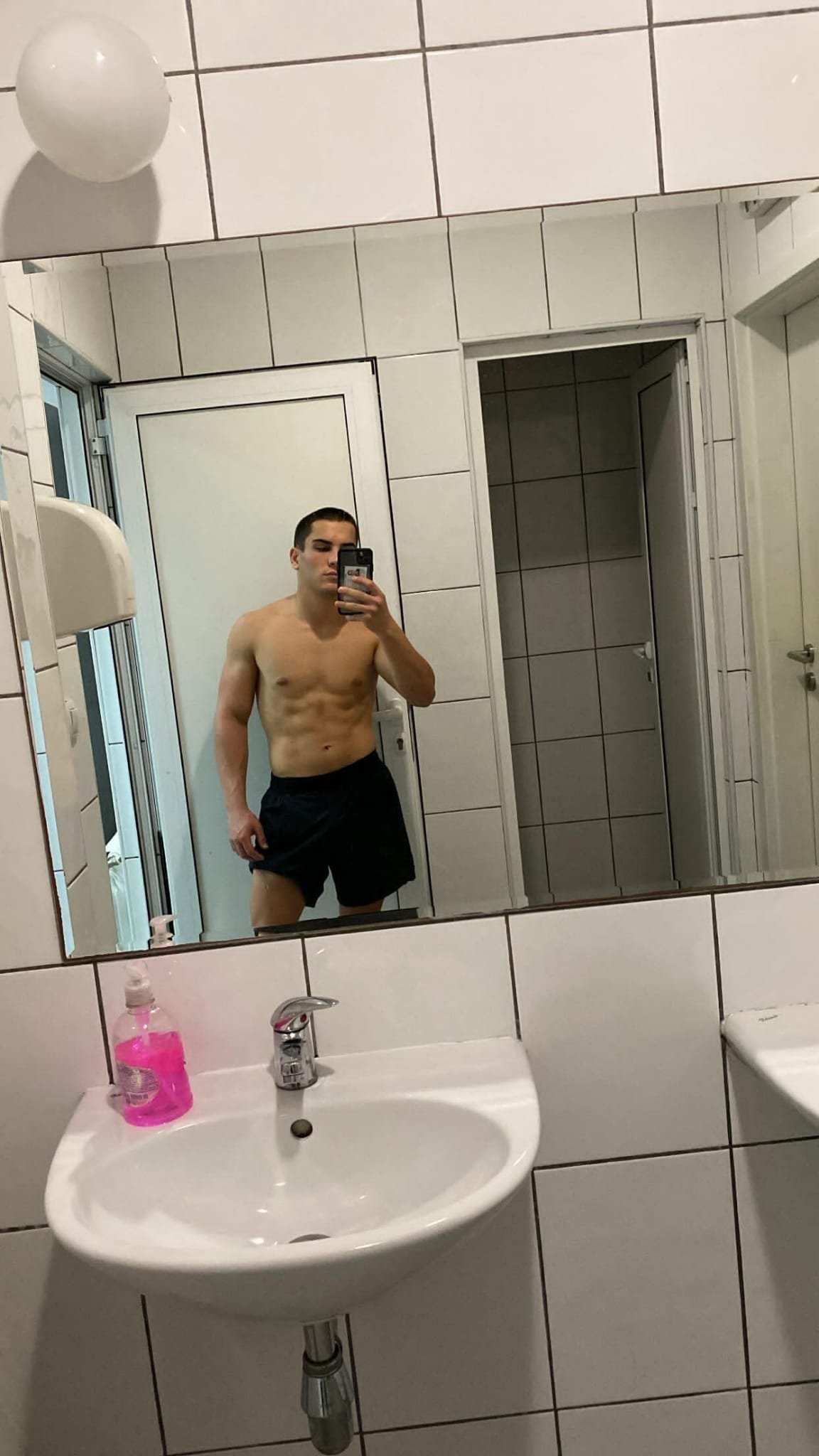 A shirtless young man taking a mirror selfie in a bathroom, standing next to a sink with pink soap, with a tiled wall and a shower area in the background.