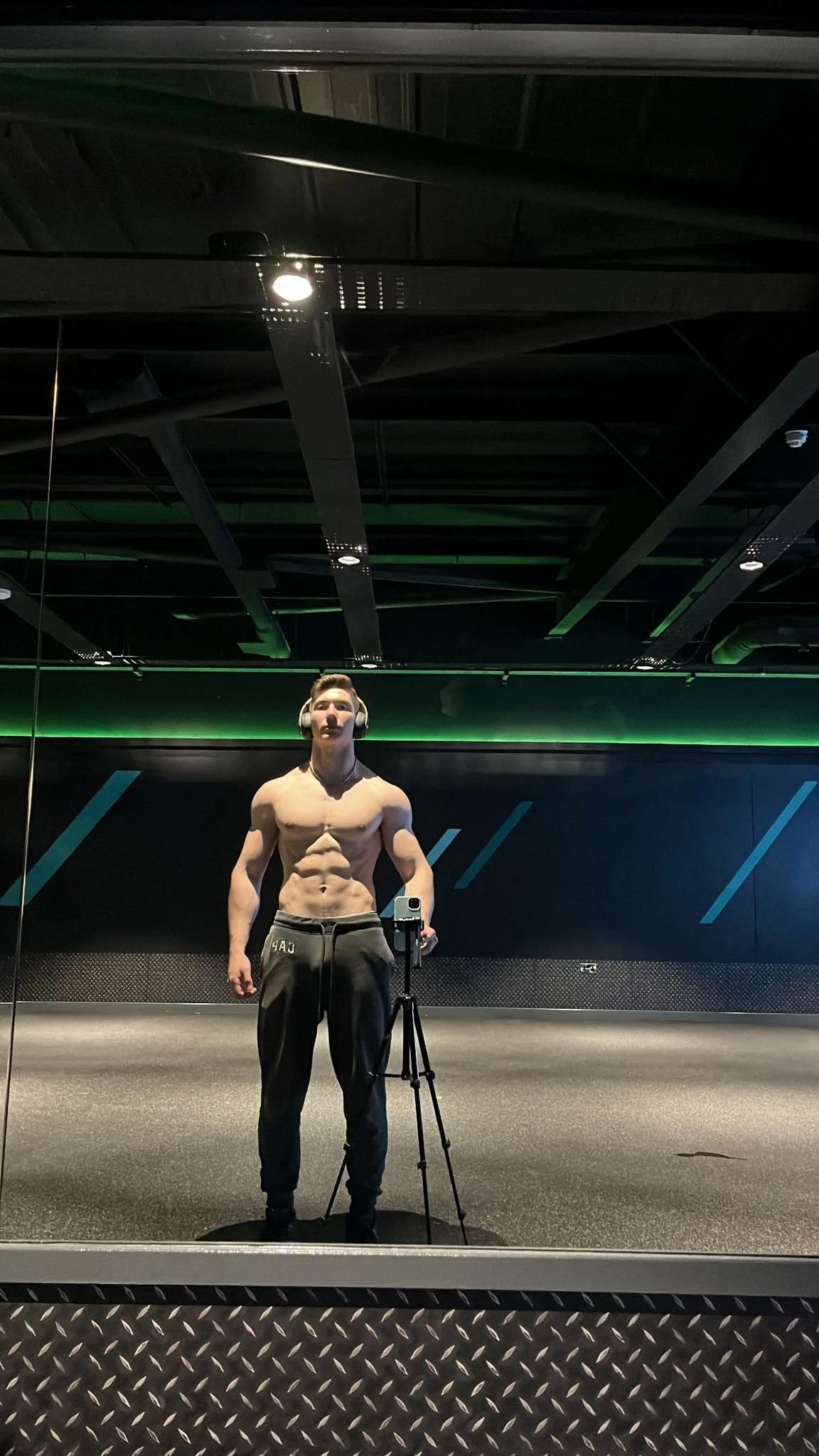 A shirtless man wearing headphones and sweatpants taking a selfie in a gym with a large mirror.