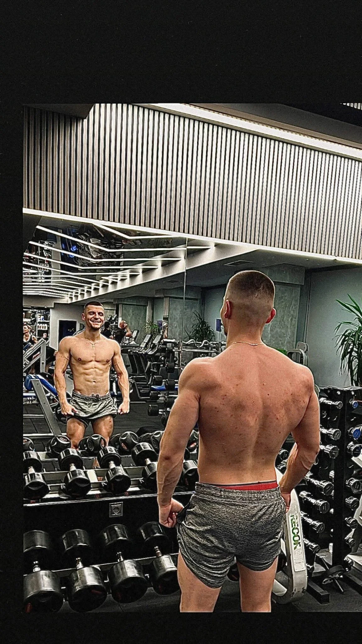 A muscular shirtless man looking at himself in a gym mirror, smiling, with dumbbells and fitness equipment in the background.