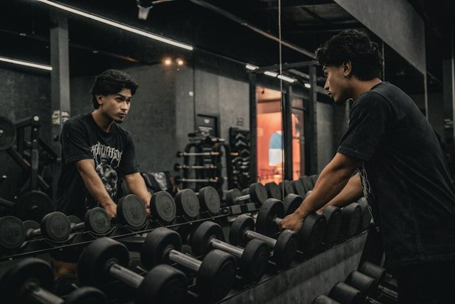 A young man with dark hair lifting dumbbells in front of a mirror at a gym, wearing a black T-shirt.